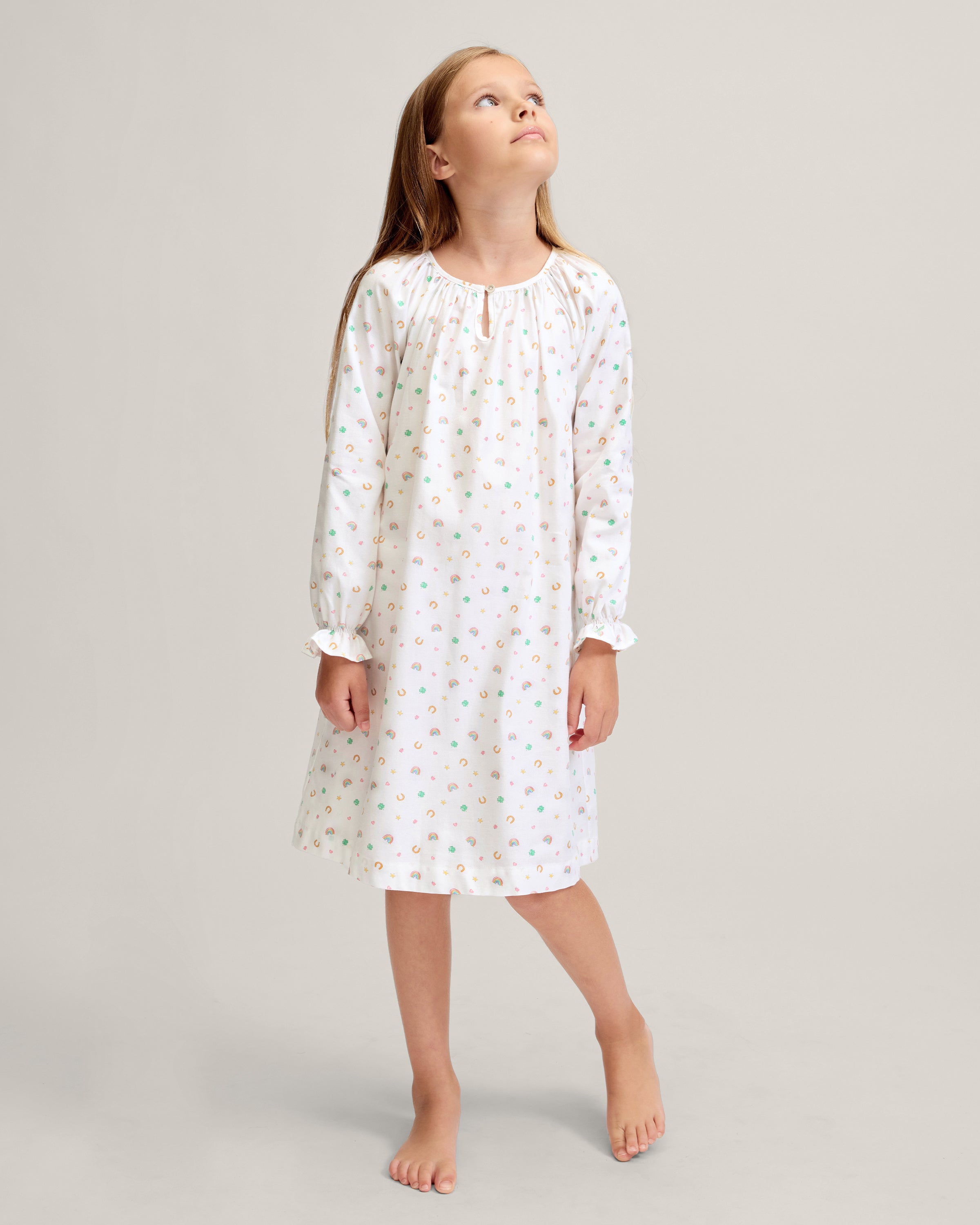 A young girl stands barefoot on a light background, wearing the Girl's Twill Delphine Nightgown in Good Luck Charms. She gazes upward thoughtfully, showcasing its long sleeves and colorful pattern.