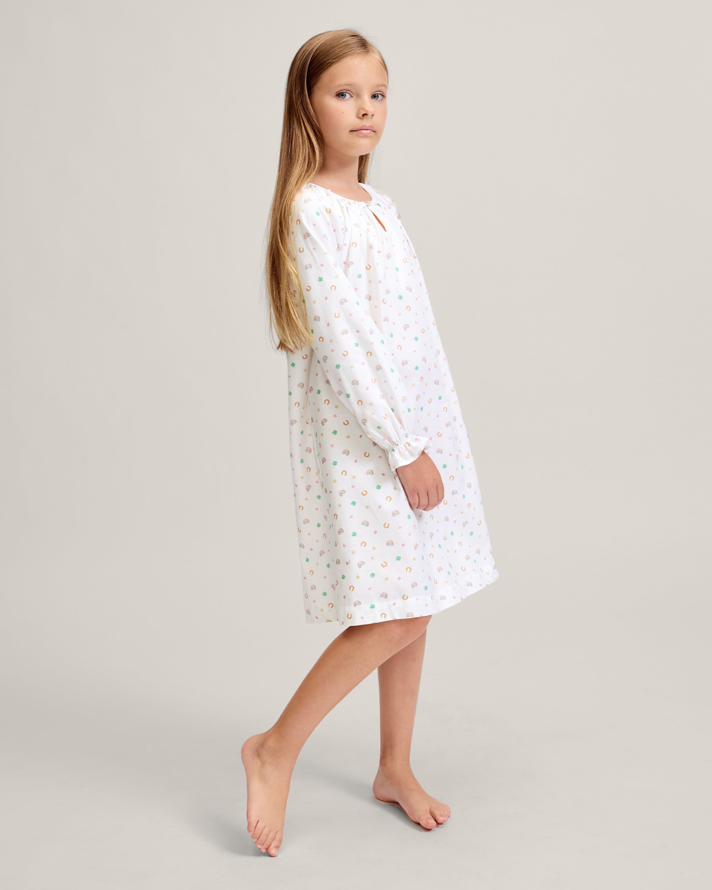A young girl stands barefoot on a light background, wearing the Girl's Twill Delphine Nightgown in Good Luck Charms. She gazes upward thoughtfully, showcasing its long sleeves and colorful pattern.