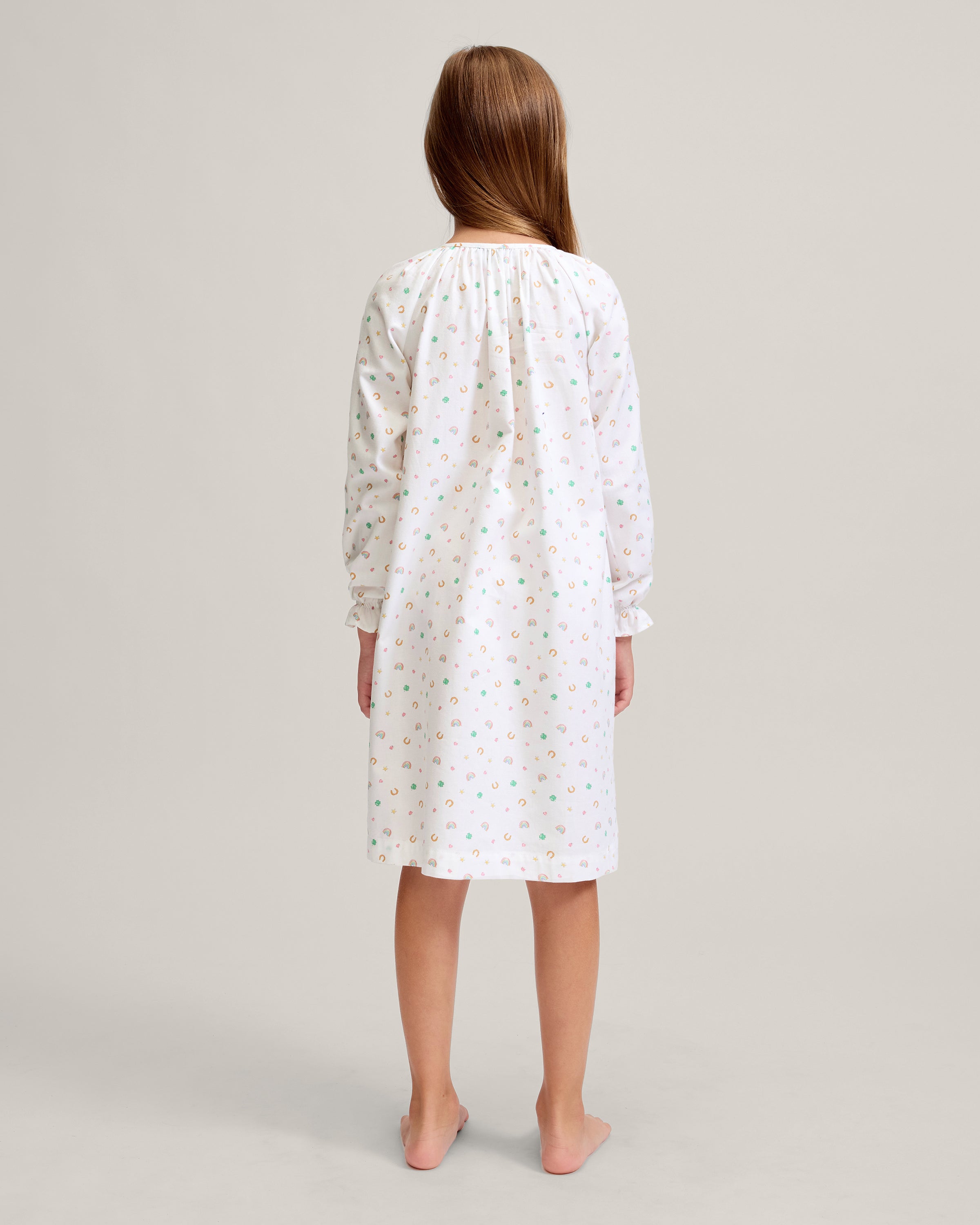 A young girl stands barefoot on a light background, wearing the Girl's Twill Delphine Nightgown in Good Luck Charms. She gazes upward thoughtfully, showcasing its long sleeves and colorful pattern.