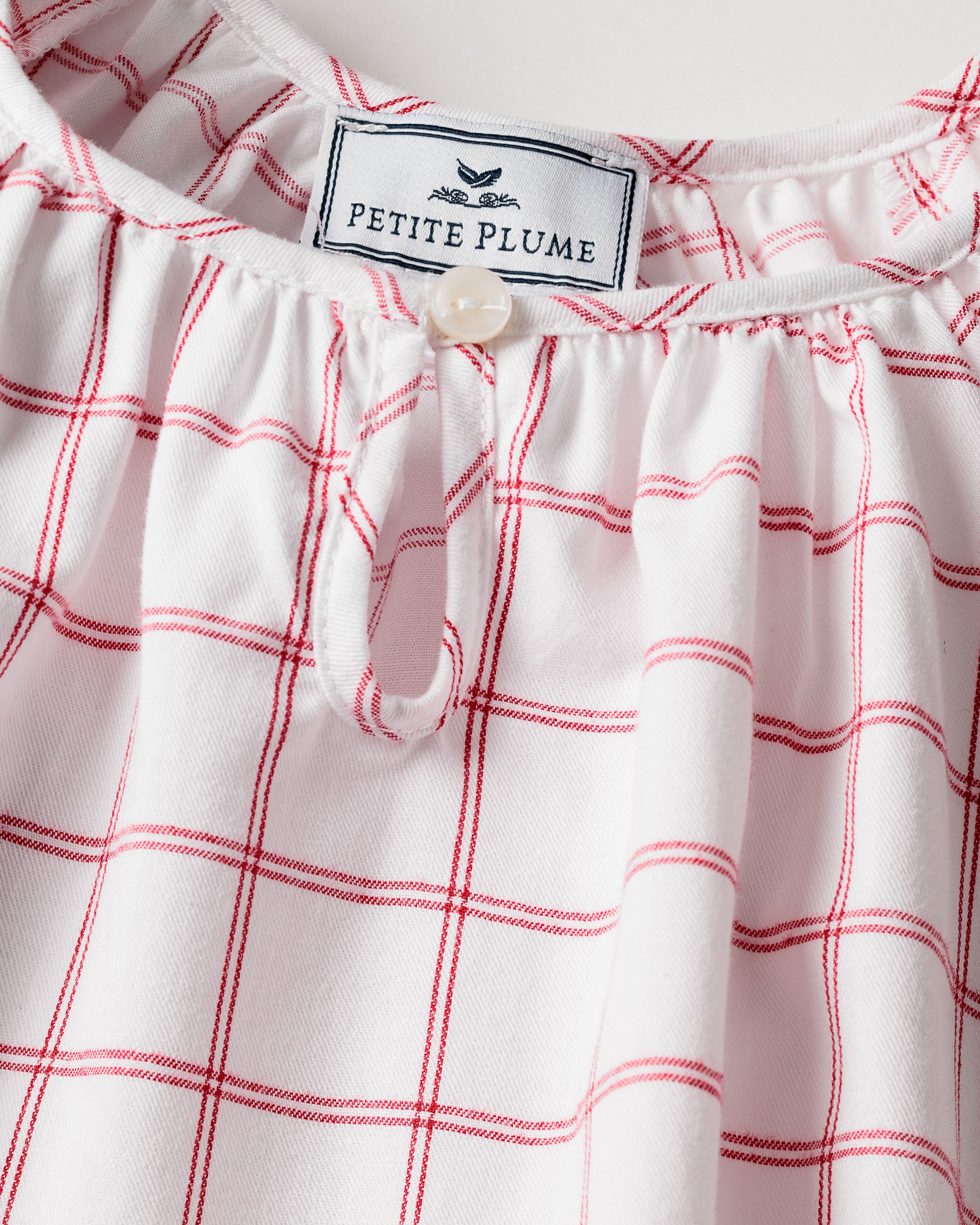 Close-up of the neckline on the Girls Twill Delphine Nightgown in Garnet Tattersall, made from flame-retardant fabric. Featuring a small white button and the Petite Plume label with a bird logo, perfect for childrens sleepwear.