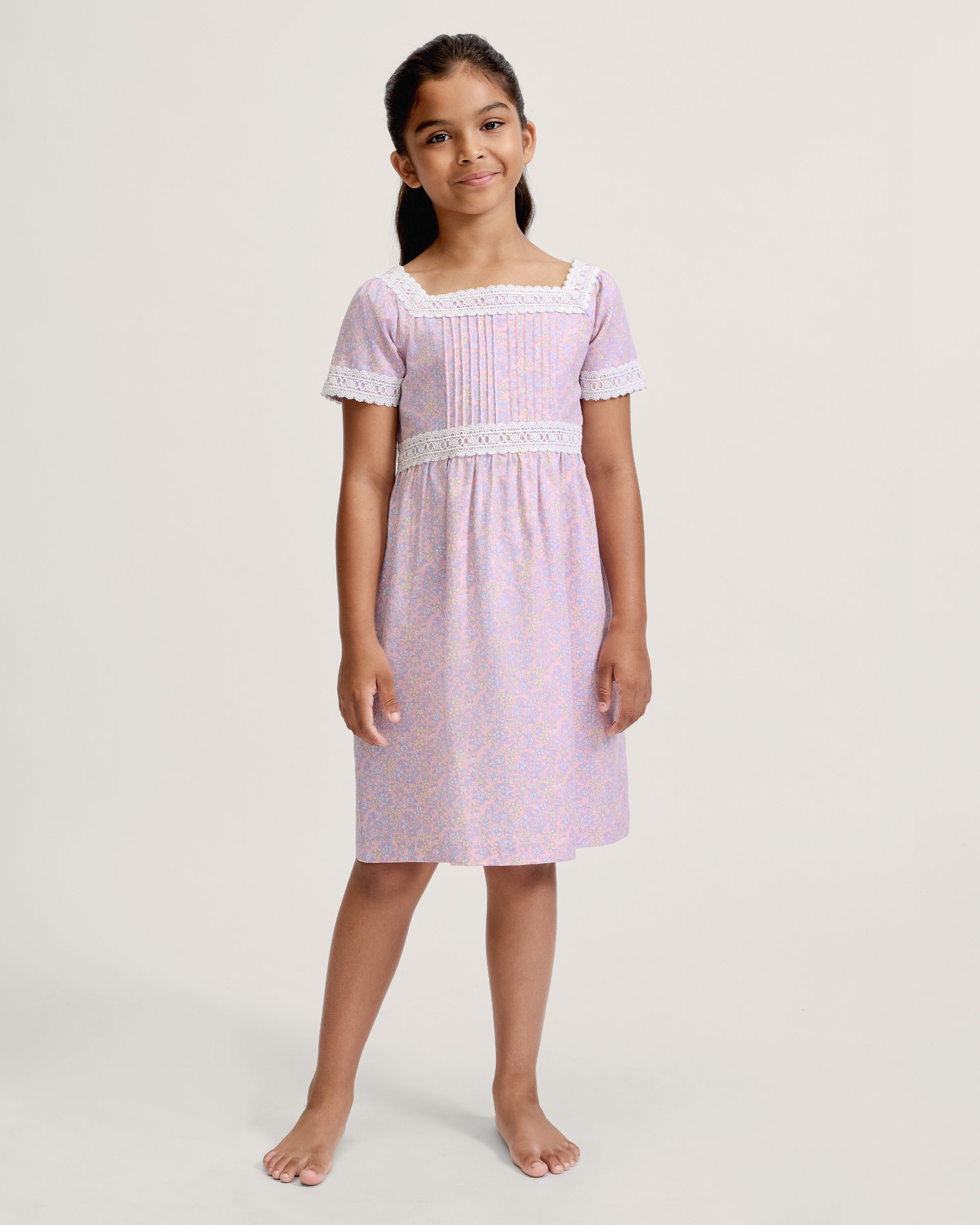 A young girl with long dark hair stands barefoot, wearing the Girl's Twill Juliette Nightgown in Fleur Blush—a light purple, knee-length dress featuring short sleeves, lace trim, and a delicate floral pattern—against a plain light background.