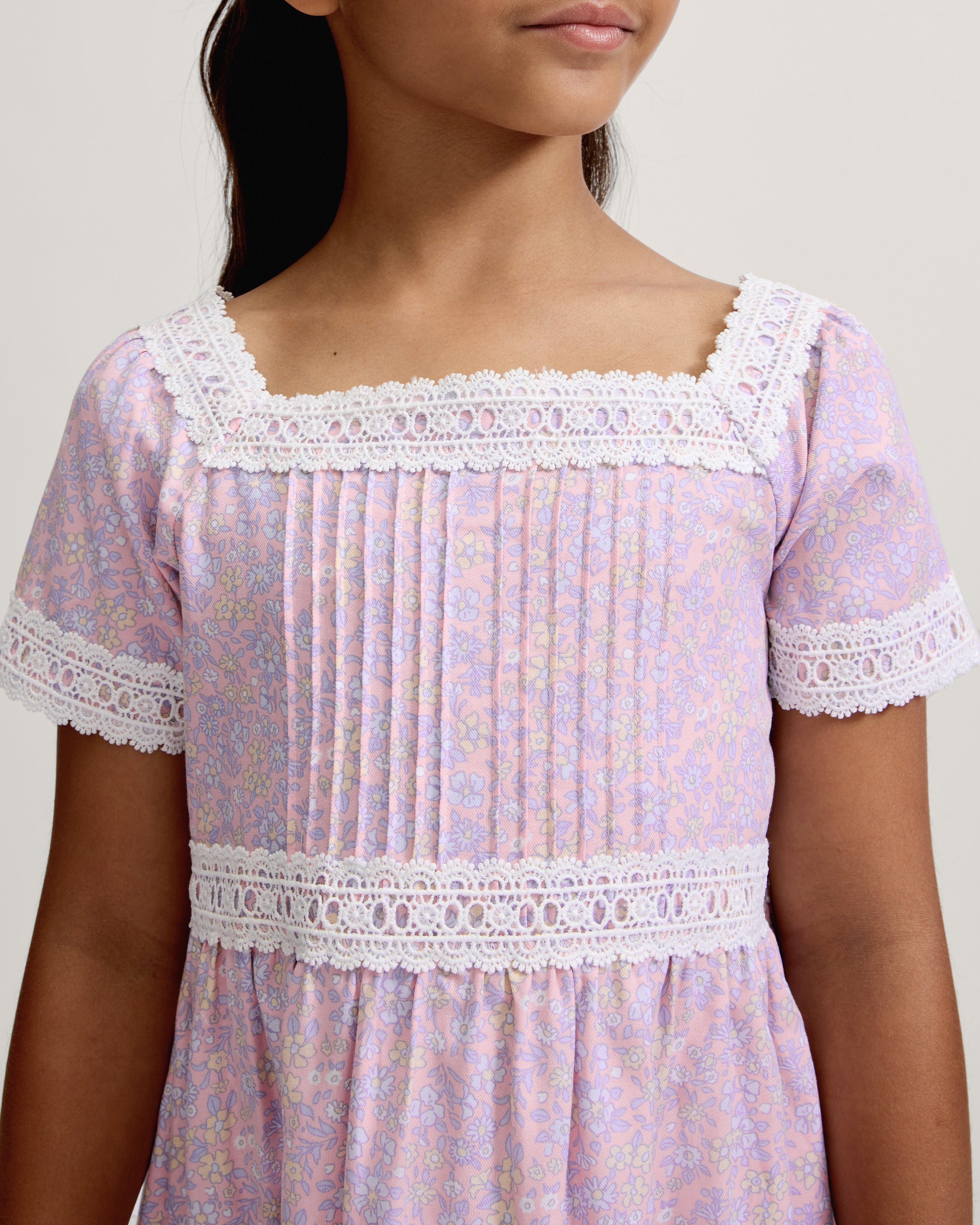 A young girl with long dark hair stands barefoot, wearing the Girl's Twill Juliette Nightgown in Fleur Blush—a light purple, knee-length dress featuring short sleeves, lace trim, and a delicate floral pattern—against a plain light background.