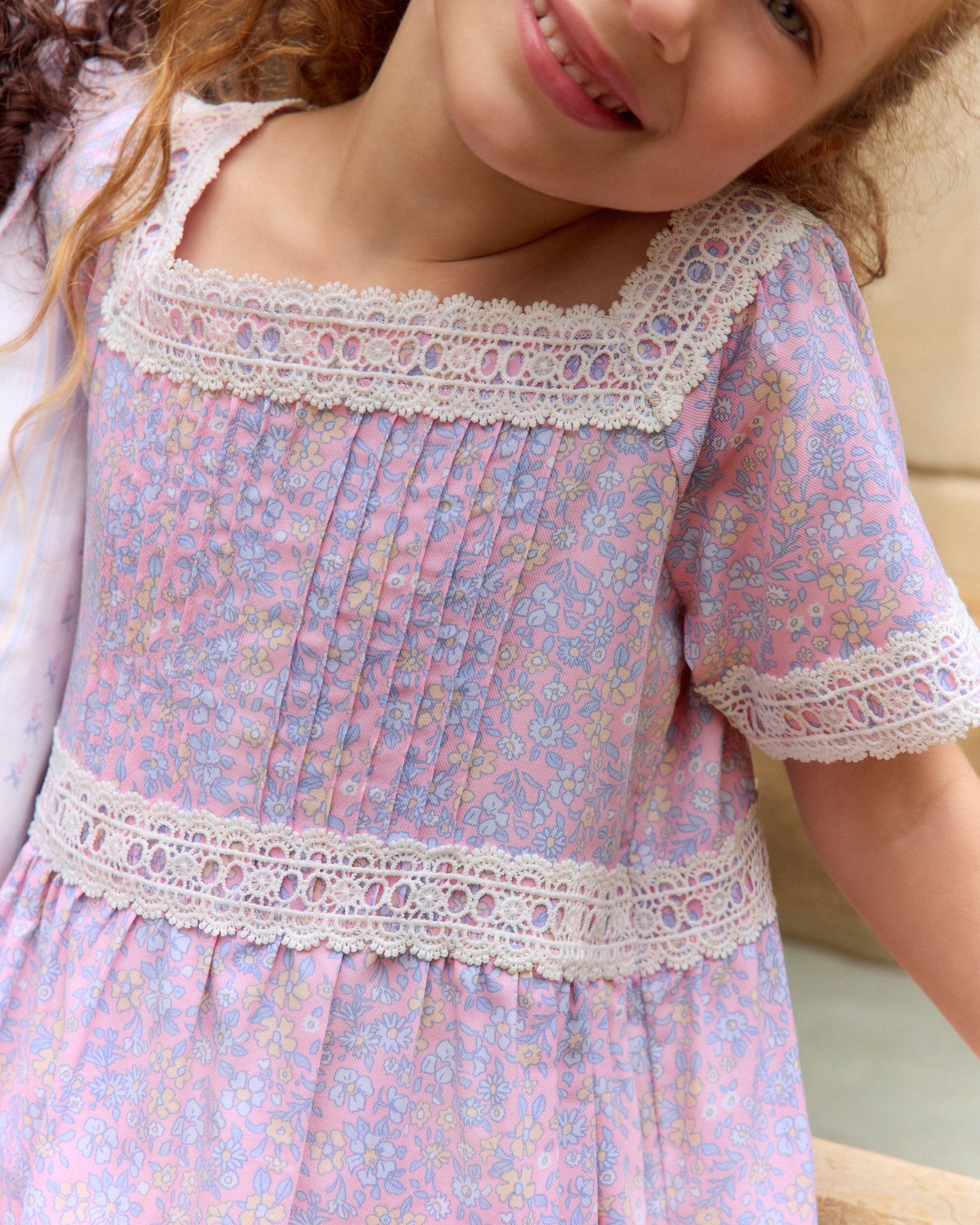 A young girl with long dark hair stands barefoot, wearing the Girl's Twill Juliette Nightgown in Fleur Blush—a light purple, knee-length dress featuring short sleeves, lace trim, and a delicate floral pattern—against a plain light background.