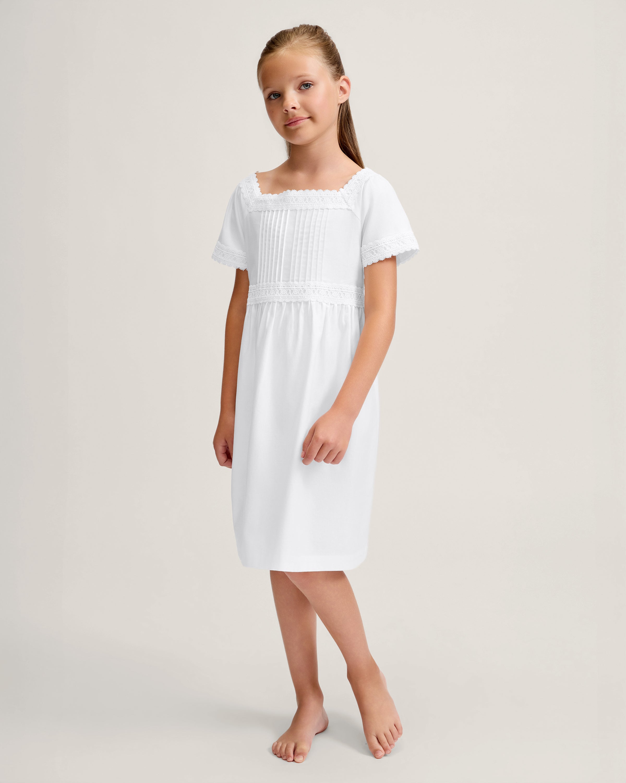 A young girl stands barefoot against a light background, wearing the Girl's Twill Juliette Nightgown in White—a knee-length piece with short sleeves, lace trim, and pintuck details. She gazes gently to the side.