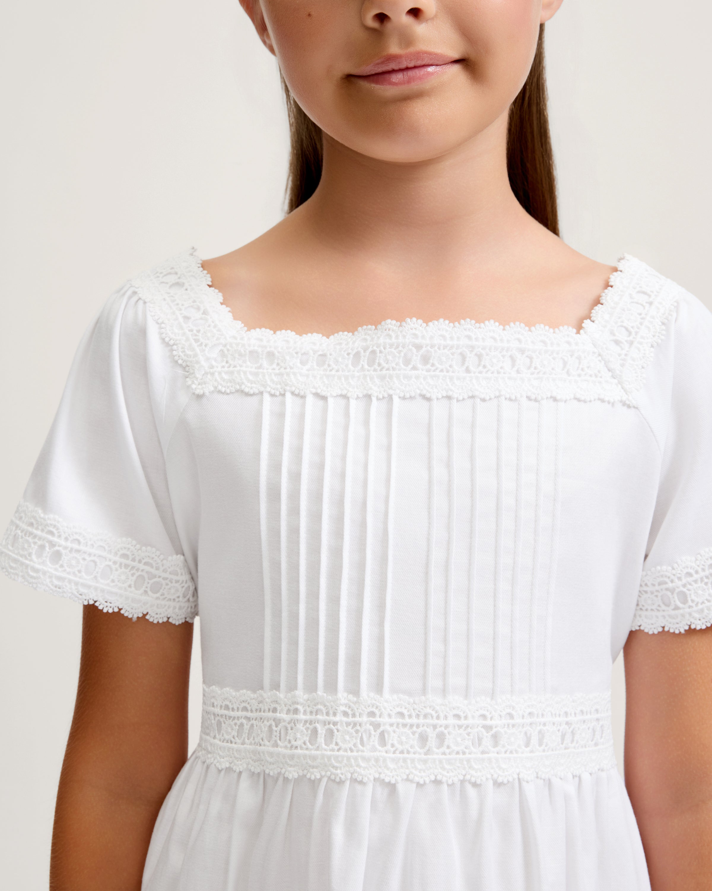 A young girl stands barefoot against a light background, wearing the Girl's Twill Juliette Nightgown in White—a knee-length piece with short sleeves, lace trim, and pintuck details. She gazes gently to the side.