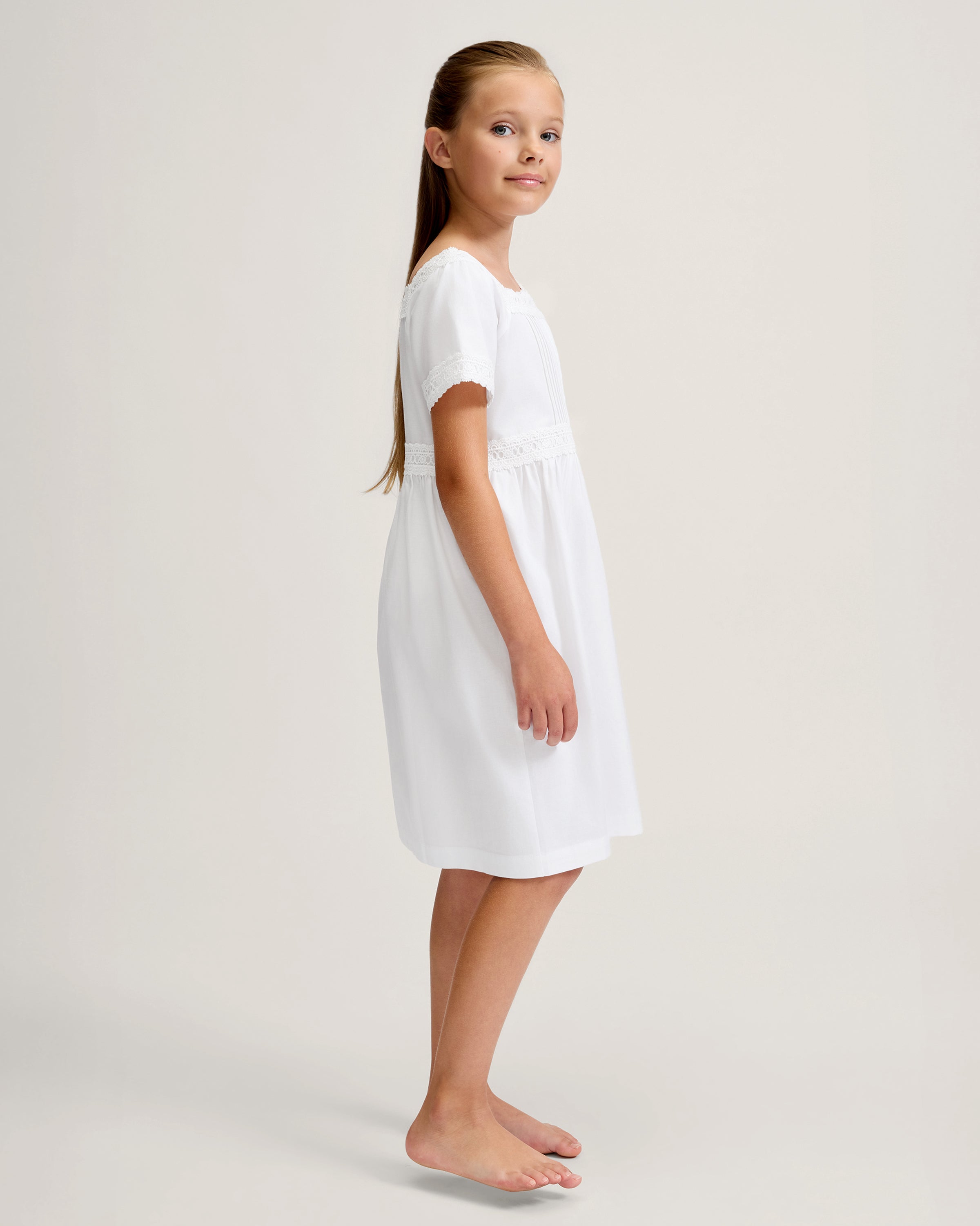 A young girl stands barefoot against a light background, wearing the Girl's Twill Juliette Nightgown in White—a knee-length piece with short sleeves, lace trim, and pintuck details. She gazes gently to the side.