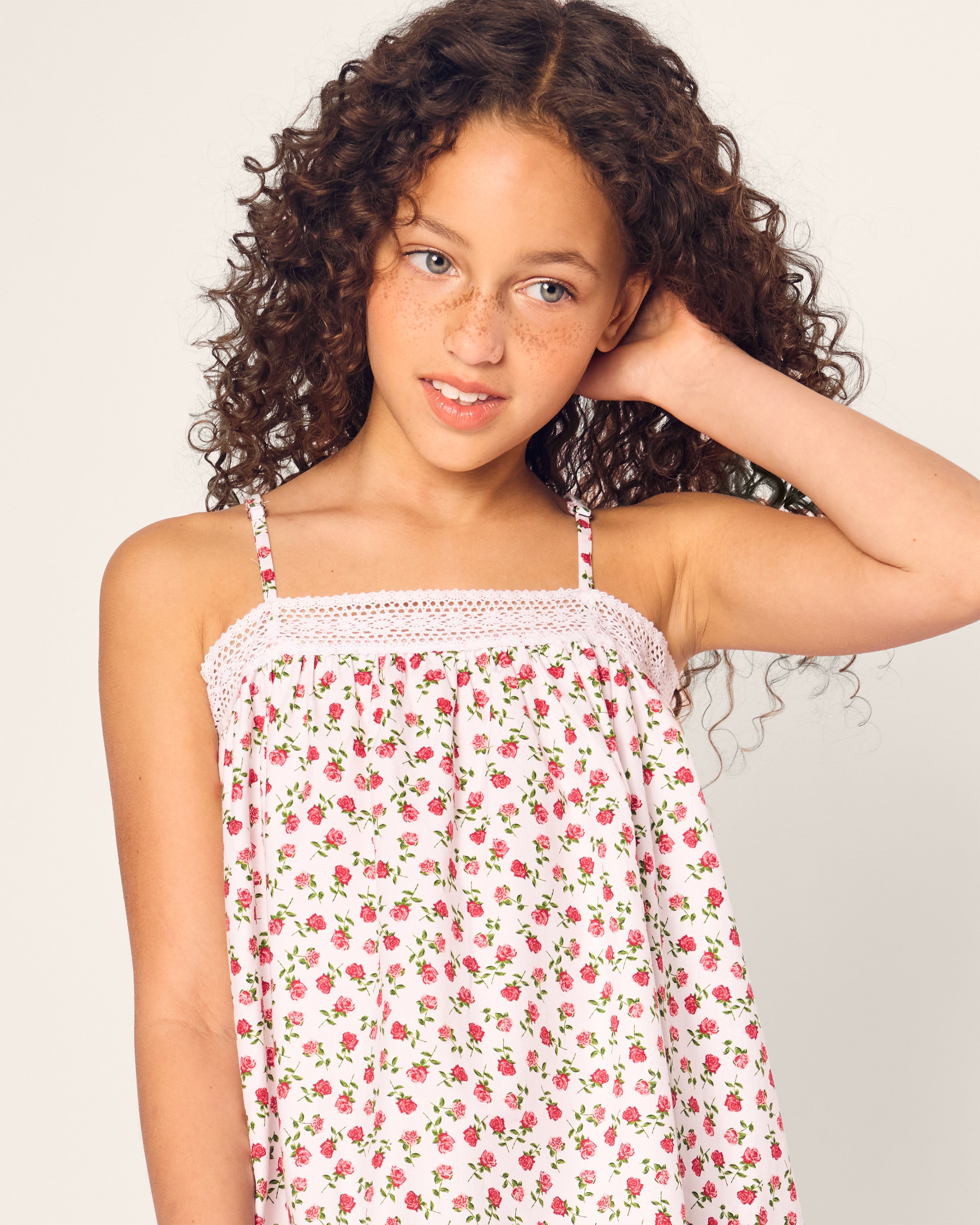 A young girl with curly hair and freckles smiles slightly, touching her hair while wearing the Petite Plume Girls Lily Nightgown in Petite Petals. She stands against a plain background.