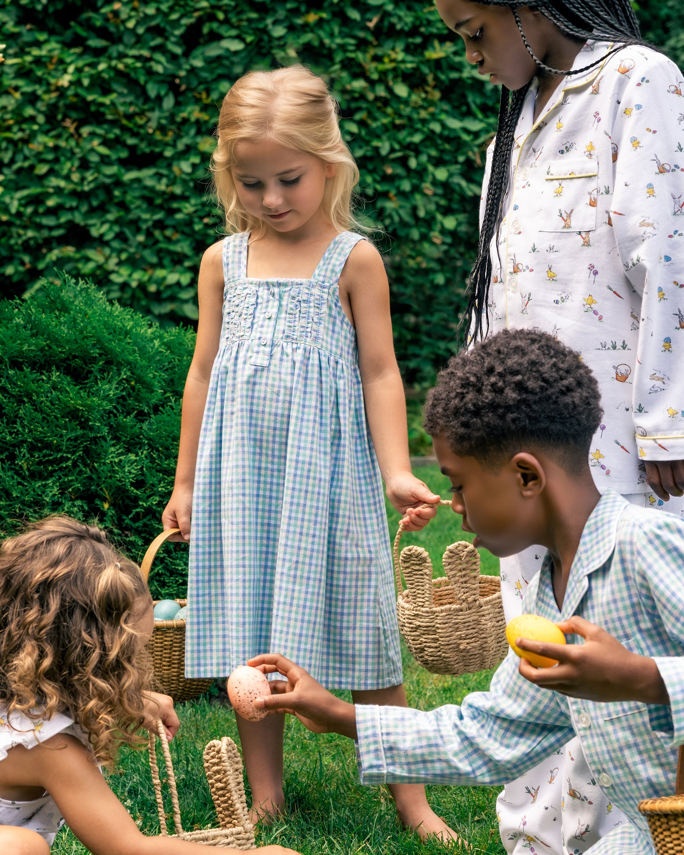 Four children play in a garden, two holding baskets of eggs. One wears flame retardant sleepwear and another twirls in the Girls Twill Charlotte Nightgown in Spring Gingham by Petite Plume. Their lively outdoor Easter egg hunt among the lush greenery brings smiles to all.
