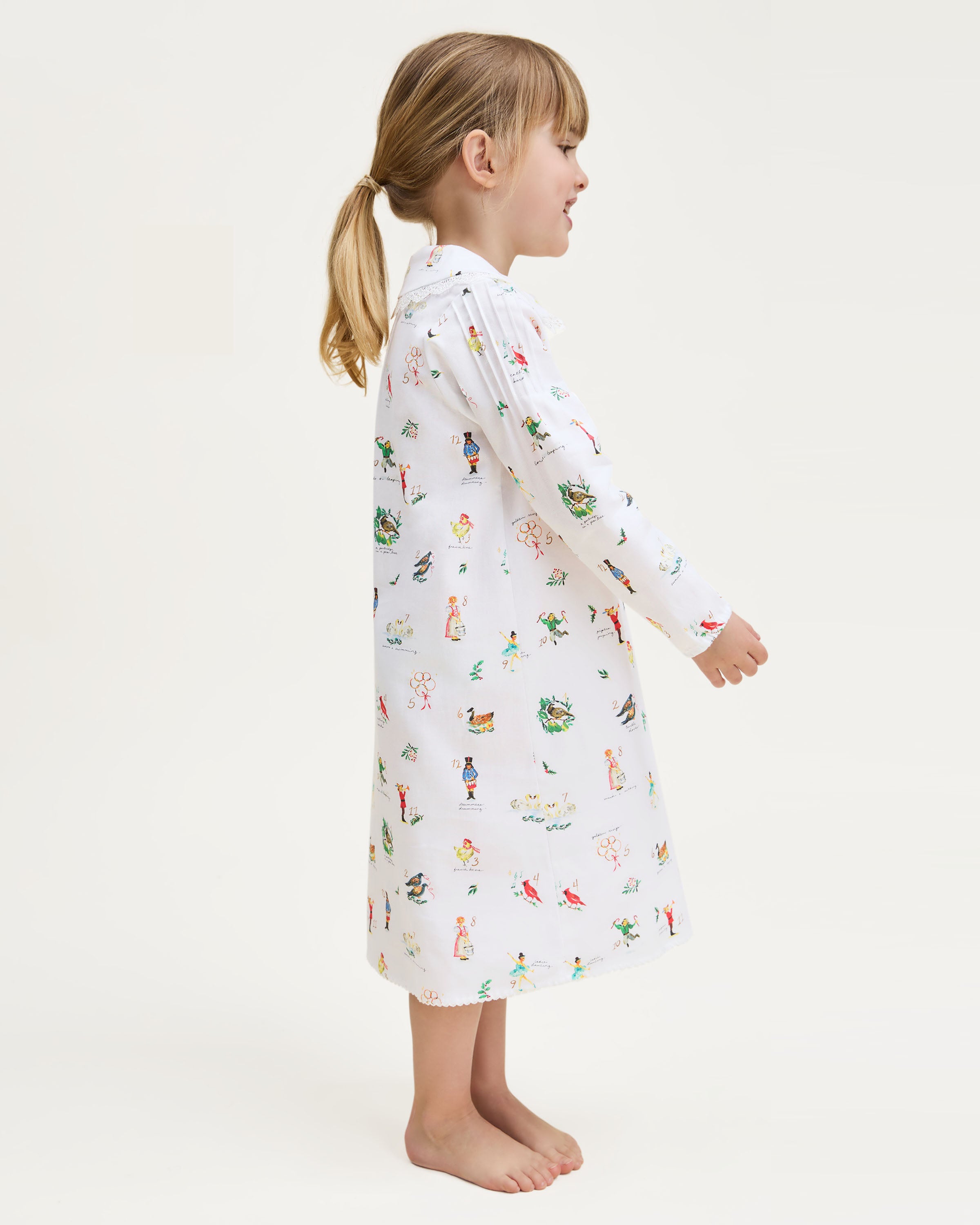 The Girl's Twill Lola Nightgown in 12 Days of Christmas is a white long-sleeve children’s dress with a Peter Pan collar and a playful pattern of colorful people, animals, and plants scattered across the fabric.