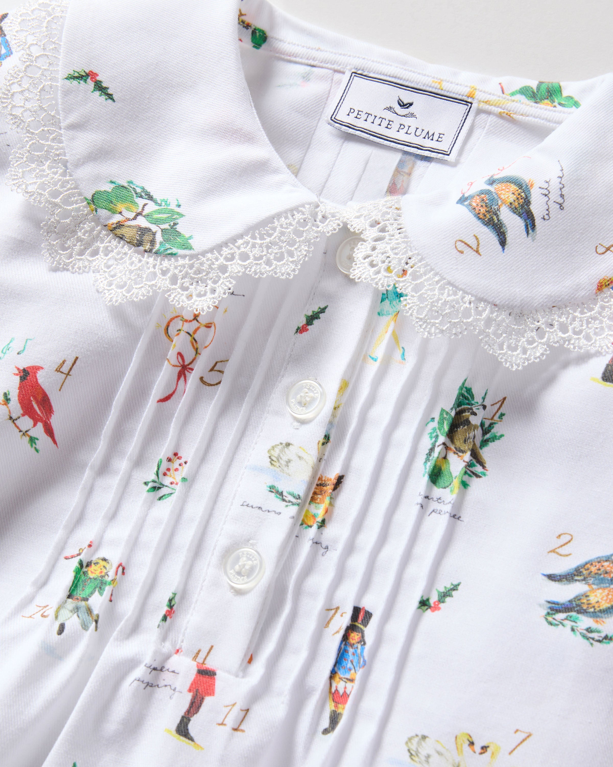 The Girl's Twill Lola Nightgown in 12 Days of Christmas is a white long-sleeve children’s dress with a Peter Pan collar and a playful pattern of colorful people, animals, and plants scattered across the fabric.