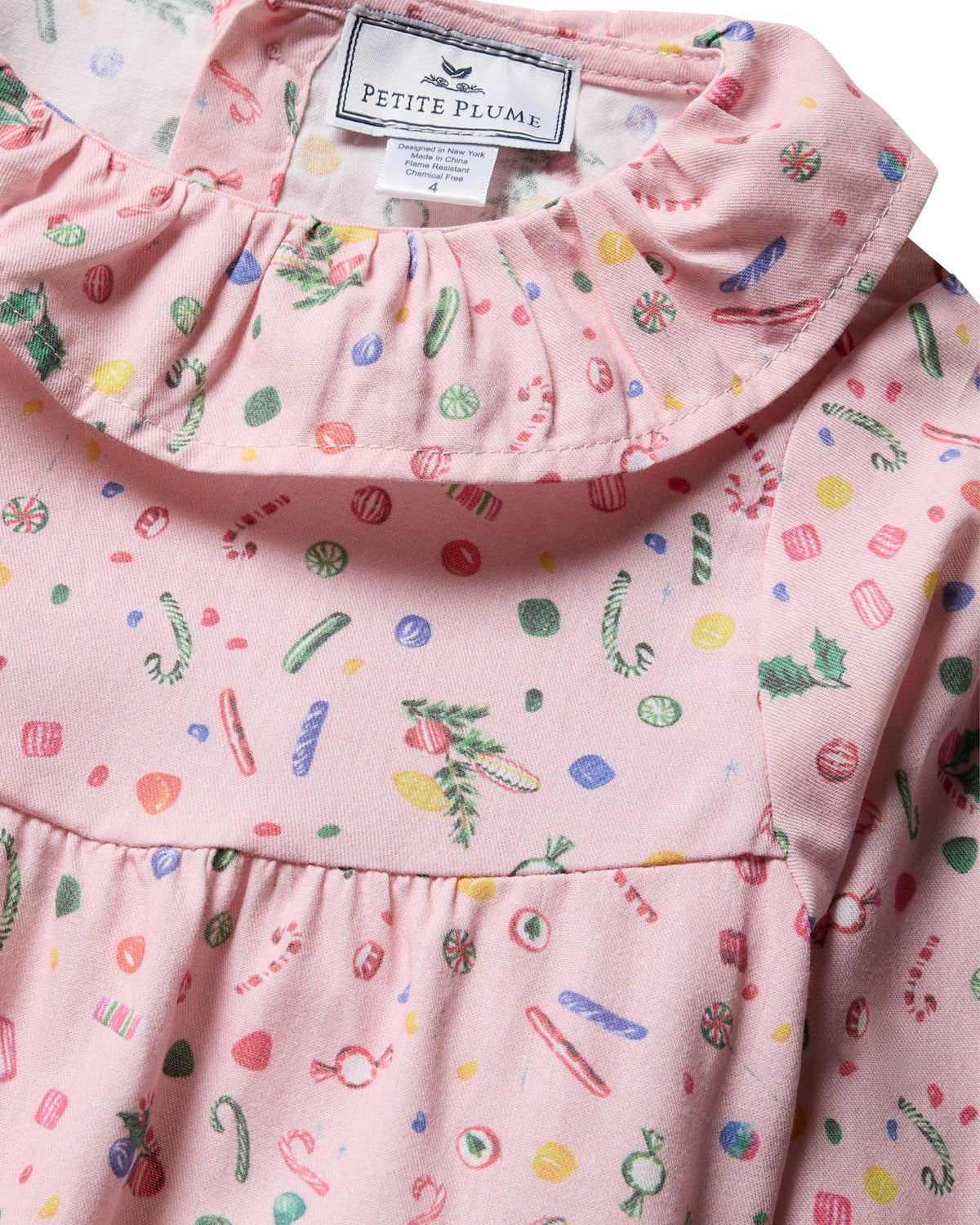 Close-up of a pink, flame-retardant fabric featuring holiday-themed candies like candy canes and peppermints. Designed as childrens sleepwear, the Petite Plume Girls Twill Scarlett Nightgown in Vintage Sweets has a gathered neckline.