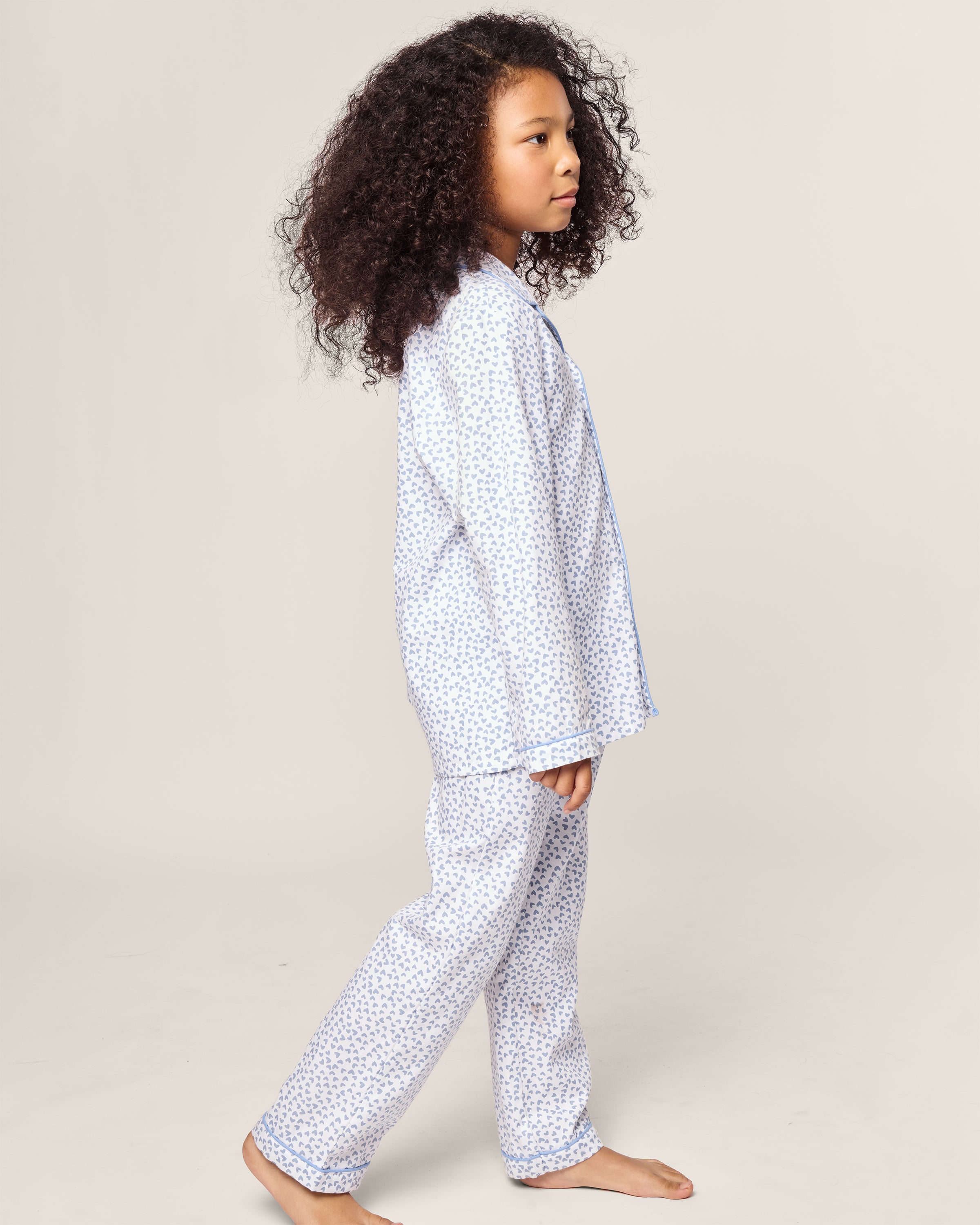 A young child with curly hair stands aside, wearing the Petite Plume Kids Twill Pajama in Bluehearts. The cozy yarn-dyed cotton set features a subtle blue floral pattern against a soft, light backdrop, enhancing the peaceful ambiance.