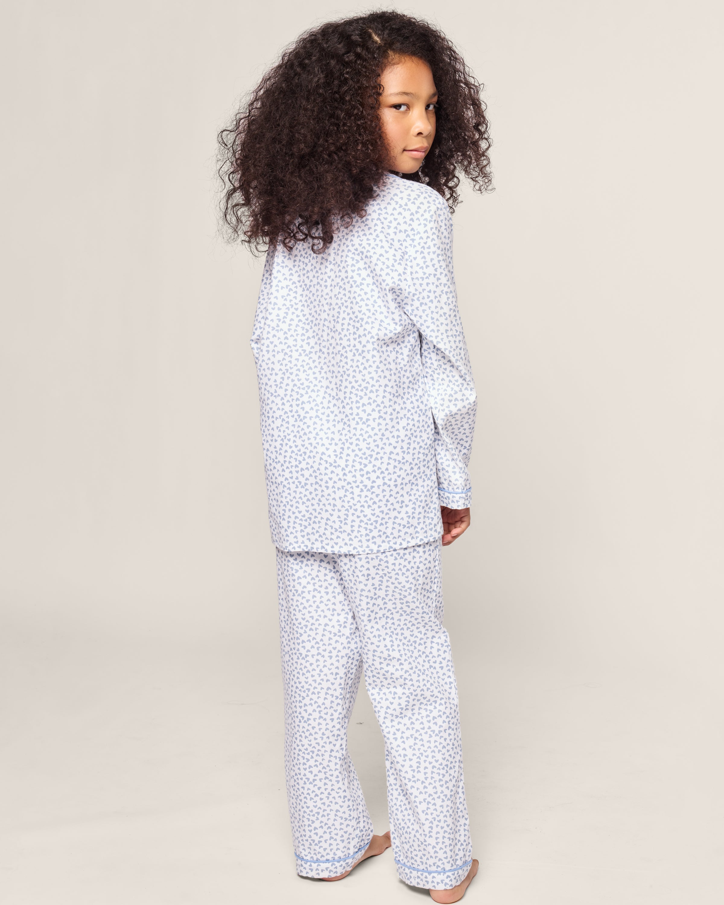 A young child with curly hair in Petite Plumes Kids Twill Pajama in Bluehearts stands barefoot on a neutral background, looking over their shoulder.