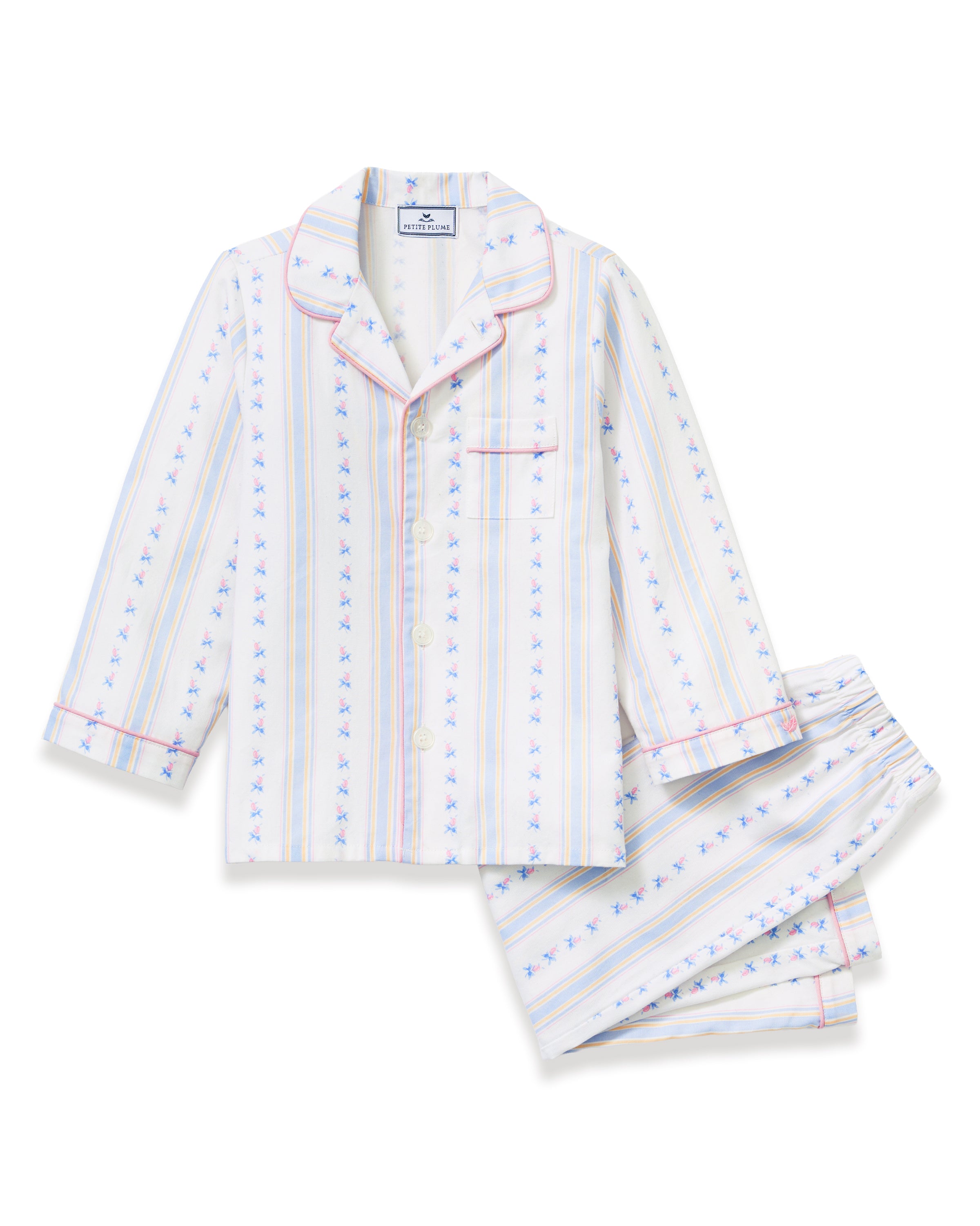 A young girl with dark hair stands barefoot on a plain background, wearing the Kid's Twill Pajama Set in Cottage Stripe—matching white pajamas with pastel stripes and a chest pocket—facing forward and smiling slightly.