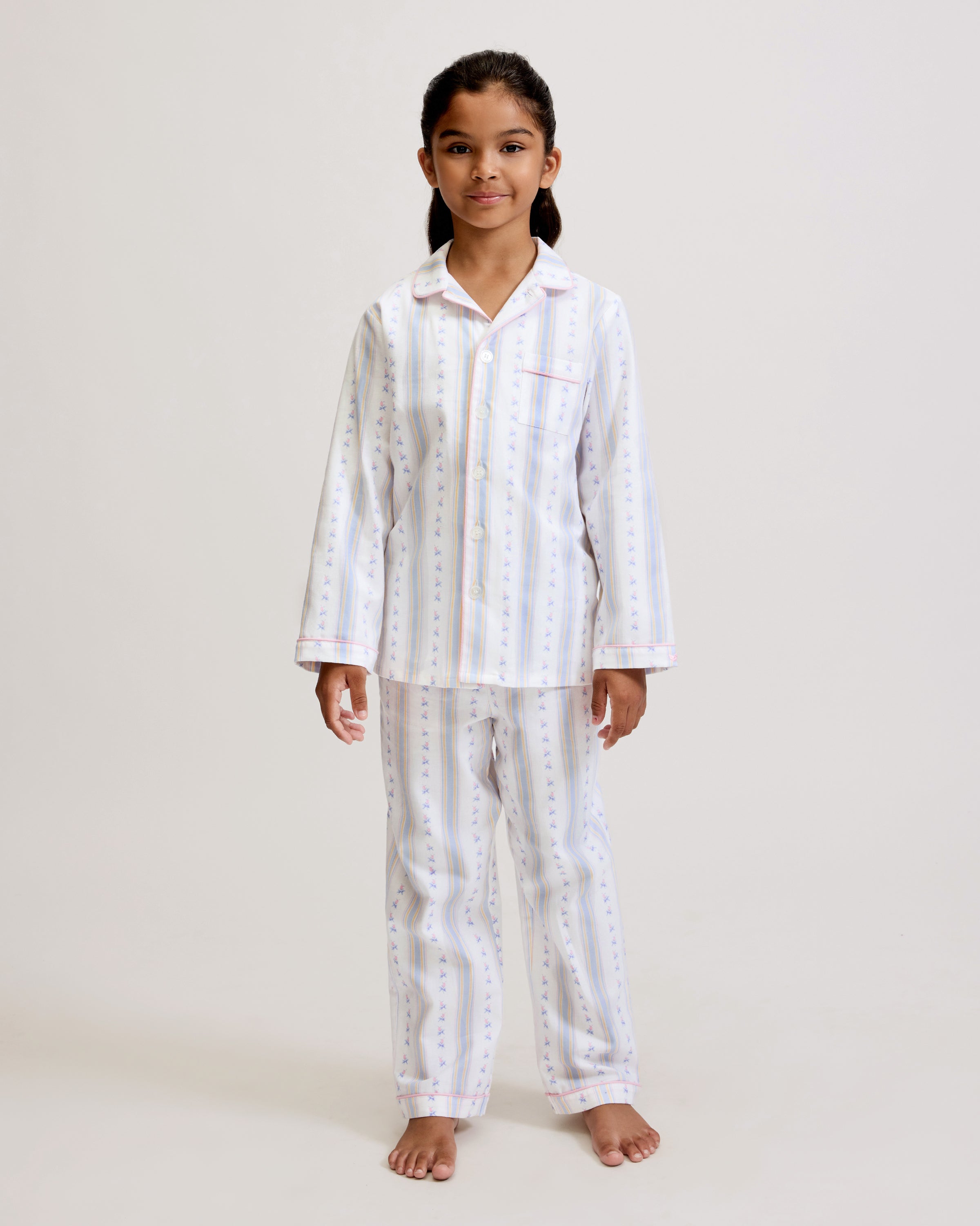 A young girl with dark hair stands barefoot on a plain background, wearing the Kid's Twill Pajama Set in Cottage Stripe—matching white pajamas with pastel stripes and a chest pocket—facing forward and smiling slightly.