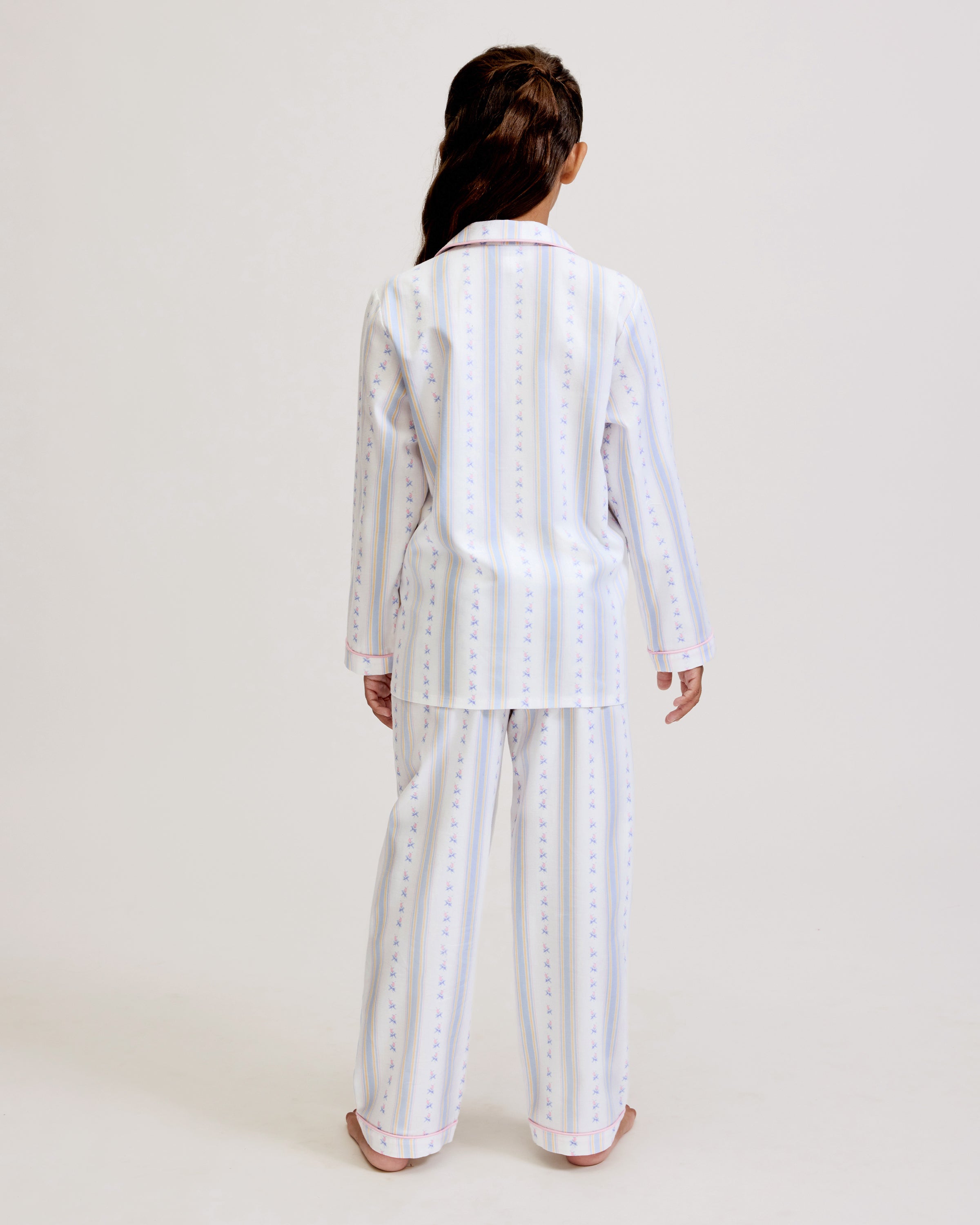 A young girl with dark hair stands barefoot on a plain background, wearing the Kid's Twill Pajama Set in Cottage Stripe—matching white pajamas with pastel stripes and a chest pocket—facing forward and smiling slightly.