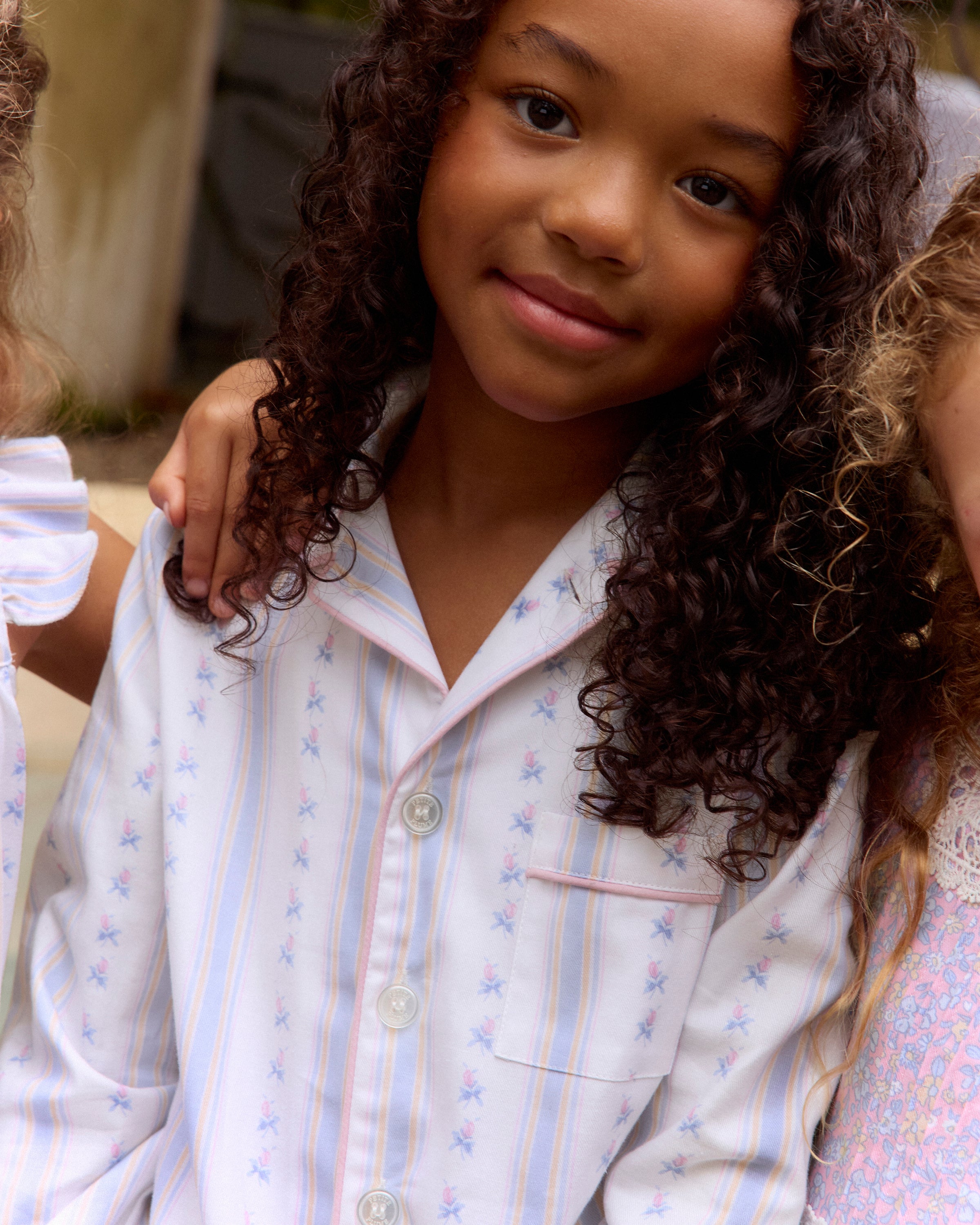 A young girl with dark hair stands barefoot on a plain background, wearing the Kid's Twill Pajama Set in Cottage Stripe—matching white pajamas with pastel stripes and a chest pocket—facing forward and smiling slightly.