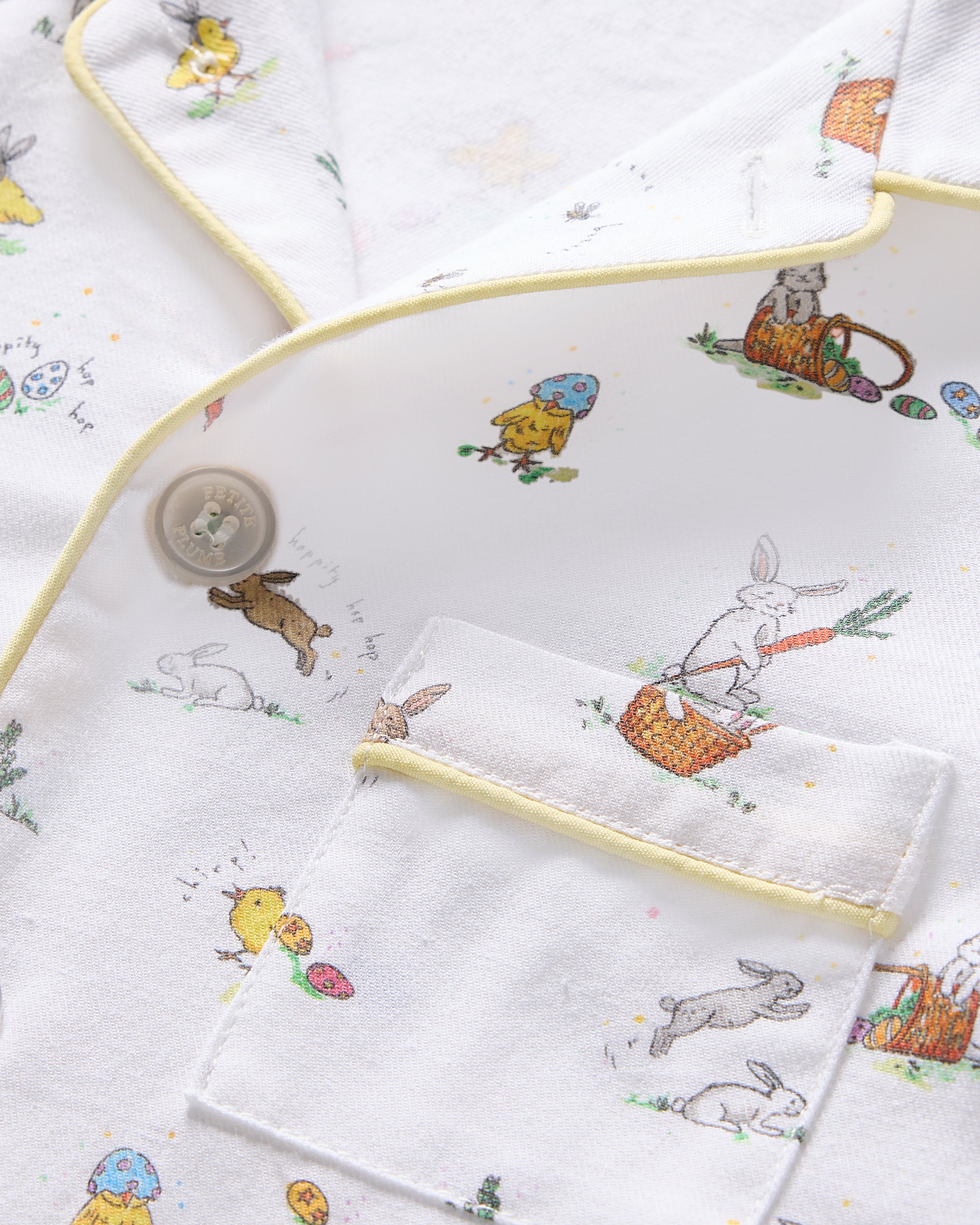 A close-up of the Kids Twill Pajama top in Easter Frolic by Petite Plume shows a white fabric with pastel piping and playful rabbits, chicks, eggs, and baskets. It features flame retardant sleepwear qualities and a pocket with button closure.