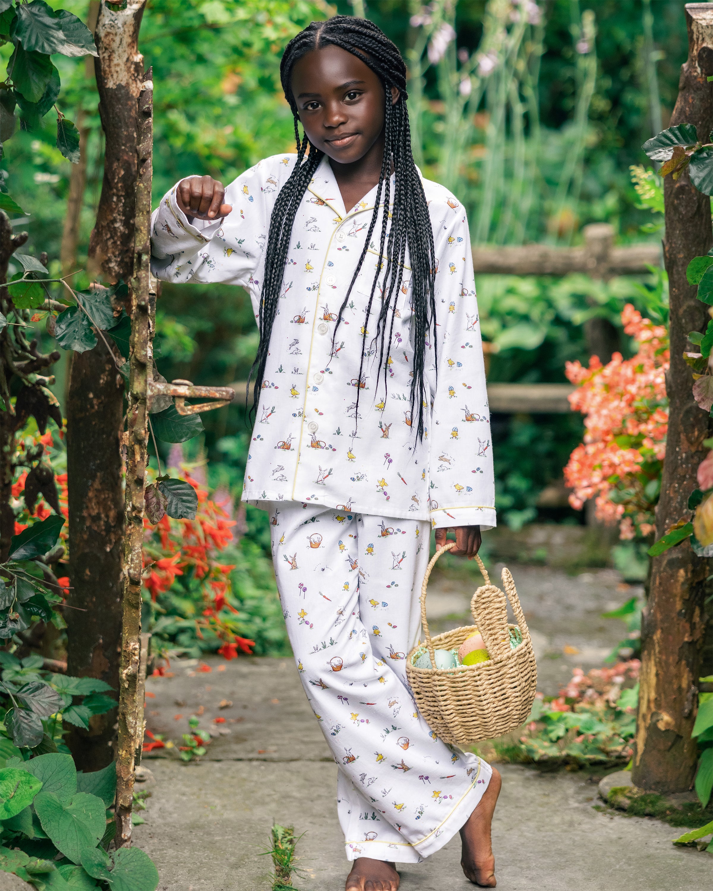 A young girl with long braided hair stands on a garden path wearing Petite Plumes Kids Twill Pajama in Easter Frolic. She smiles at the camera, holding a woven basket filled with apples, surrounded by lush greenery and flowers.