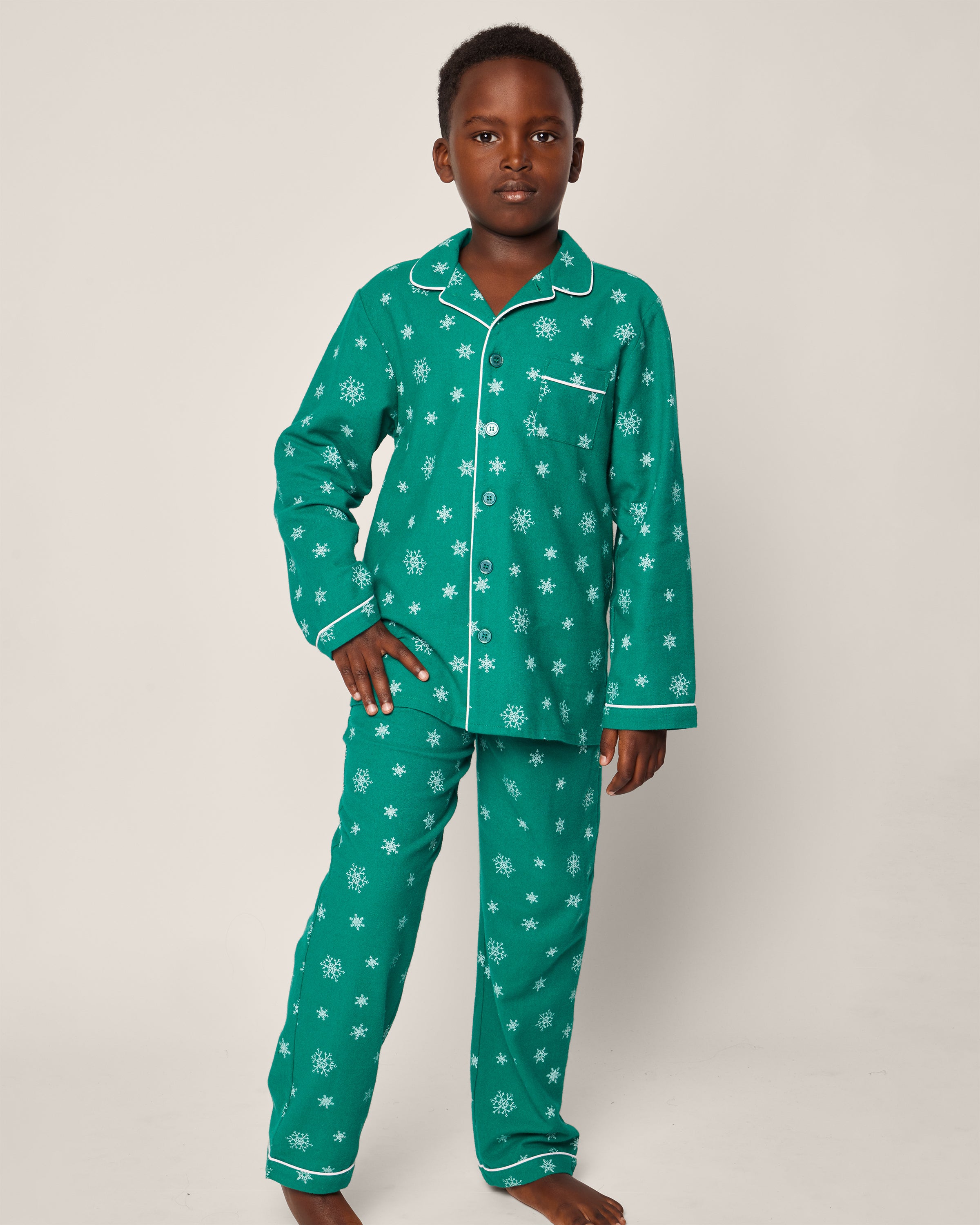 A child stands barefoot against a neutral backdrop wearing Petite Plumes Kids Flannel Pajama Set in Emerald Wonderland, featuring a green, flame-retardant design with white snowflakes, a long-sleeved collared top with a front pocket, and matching pants.