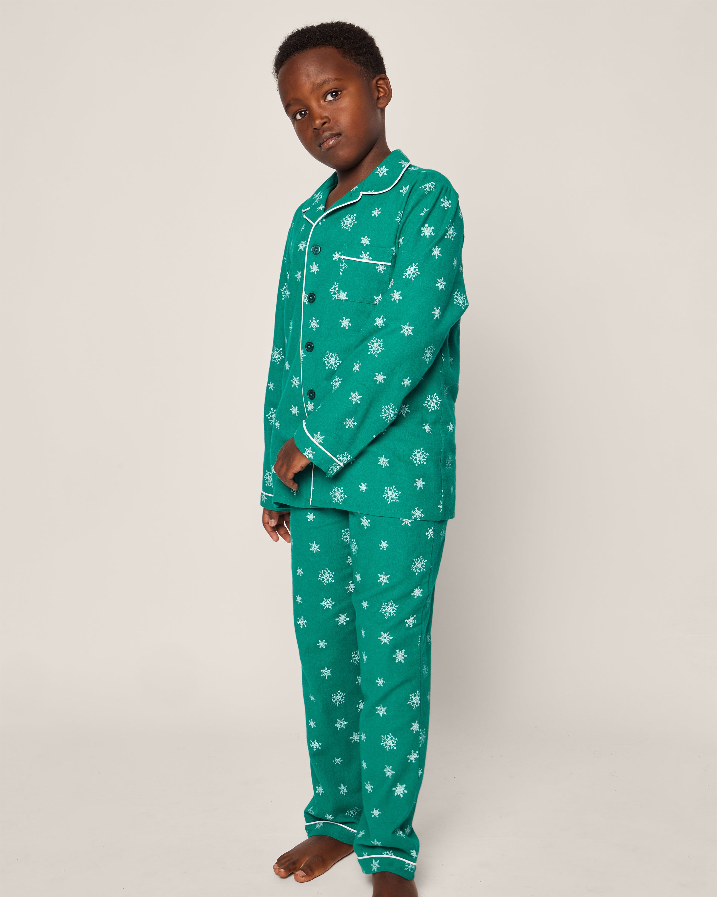 A child in Petite Plumes Kids Flannel Pajama Set in Emerald Wonderland, featuring white snowflakes on green fabric, stands barefoot against a plain backdrop. The moisture-wicking twill set has a pocketed button-up shirt with long pants. The child gazes slightly to the side.
