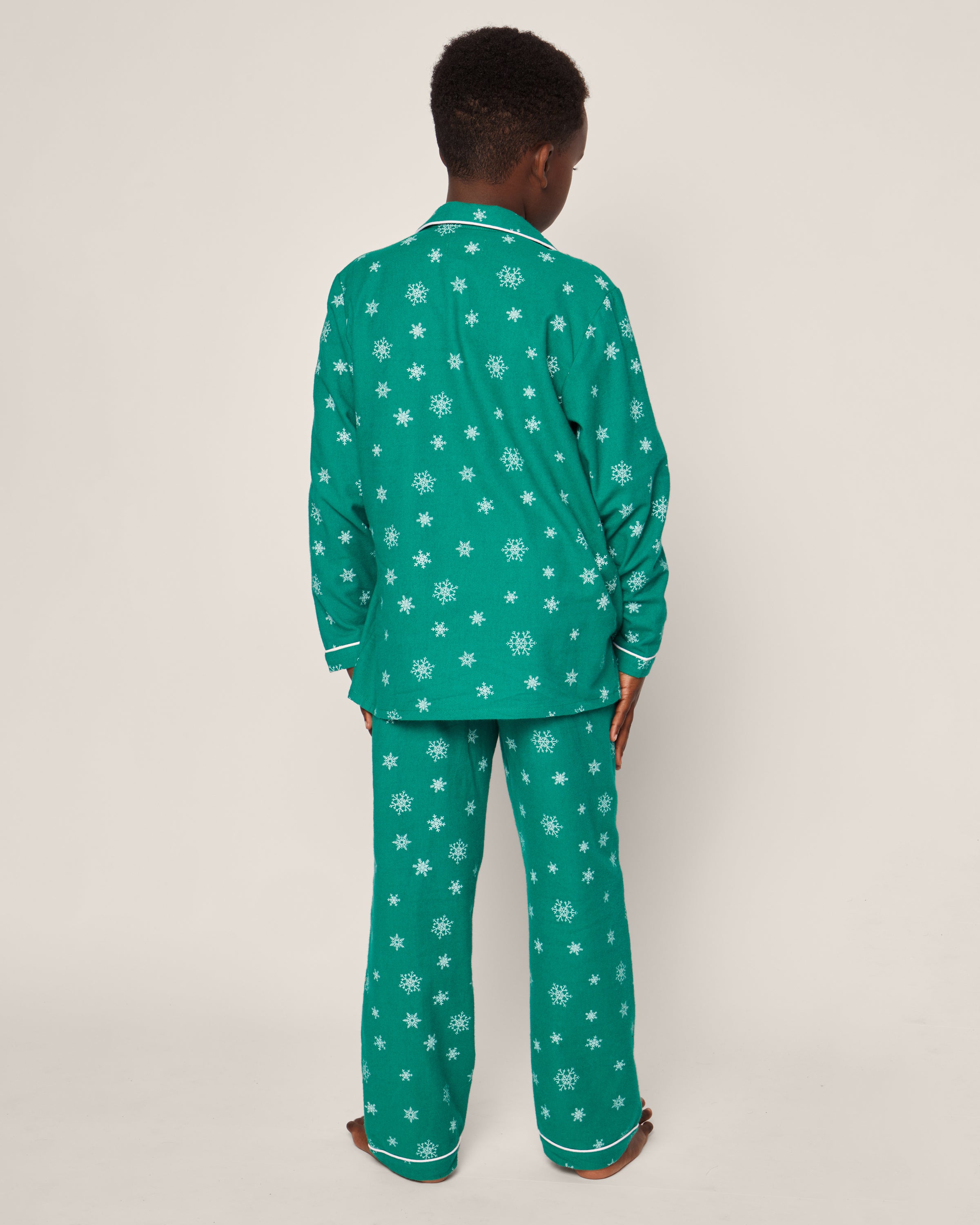 A child in Petite Plumes Kids Flannel Pajama Set in Emerald Wonderland, featuring green flame-retardant material with white snowflakes, stands facing away against a plain background.
