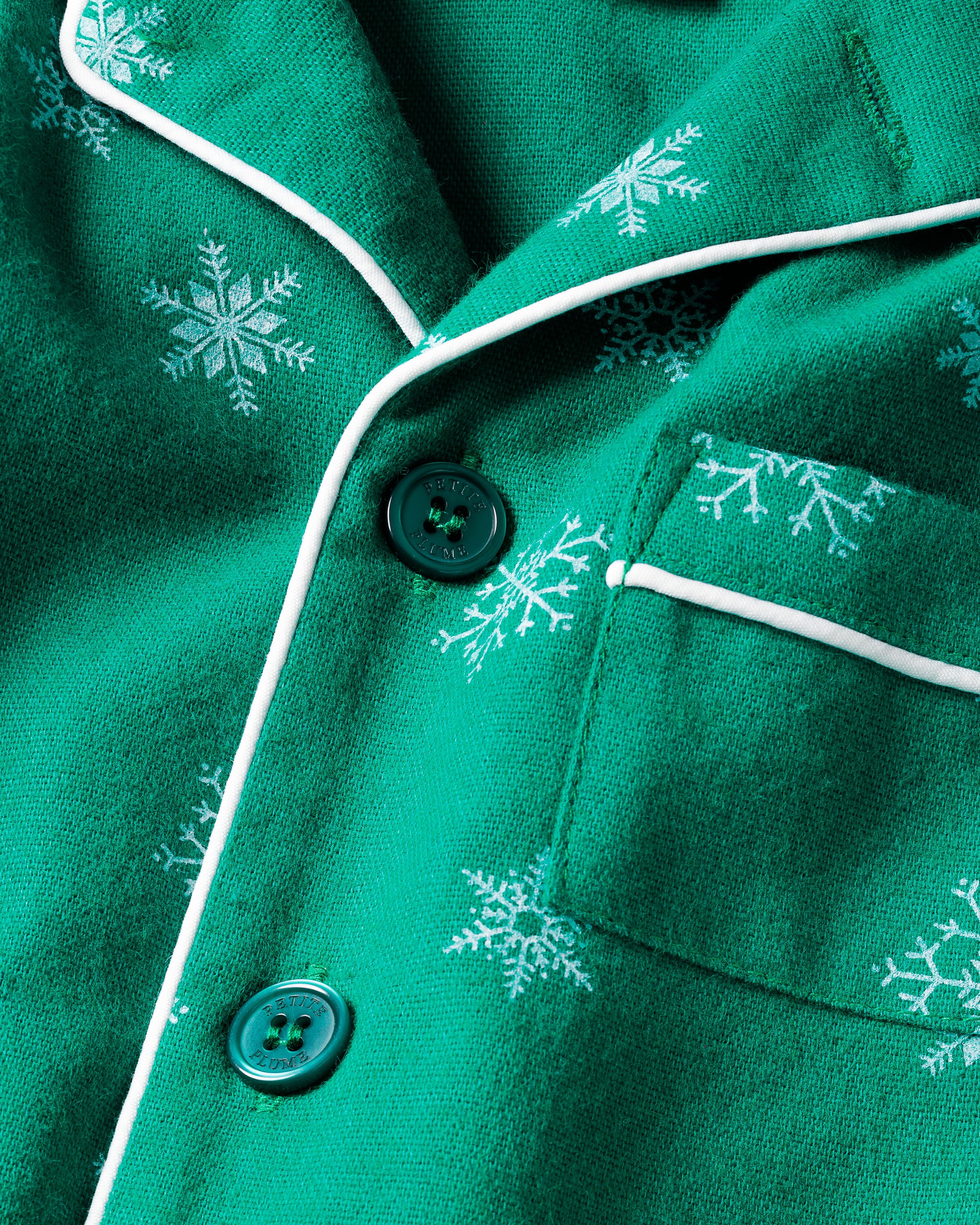 Close-up of the Kids Flannel Pajama Set in Emerald Wonderland by Petite Plume, showcasing white snowflake patterns on a green background, with green buttons and white piping near a pocket. Crafted from moisture-wicking twill.