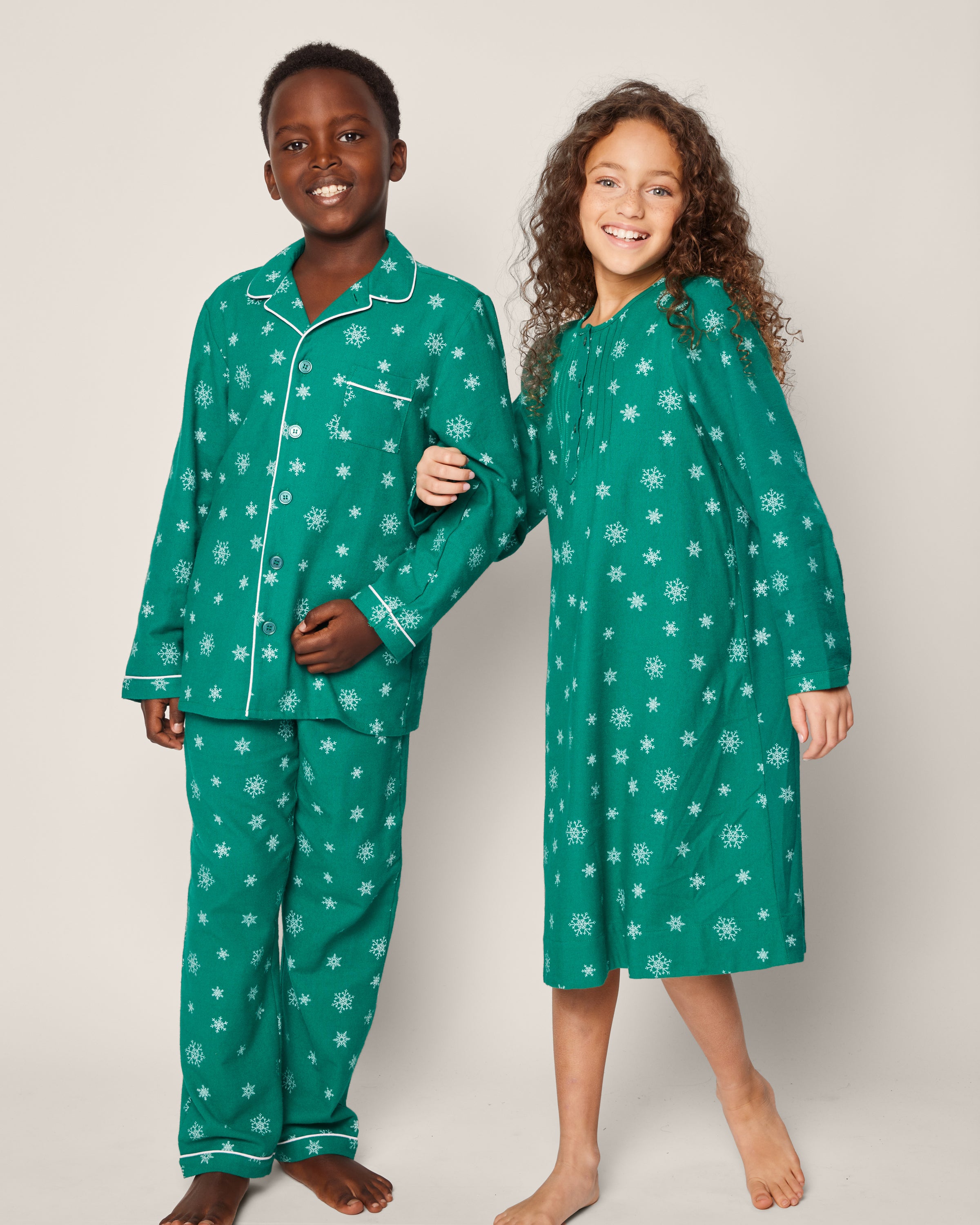 Two children beam in matching Petite Plume sleepwear: the boy sports the Kids Flannel Pajama Set in Emerald Wonderland, and the girl dons a nightgown. They are barefoot against a light background, showcasing the cozy charm of festive childrens pajamas with snowflake patterns.
