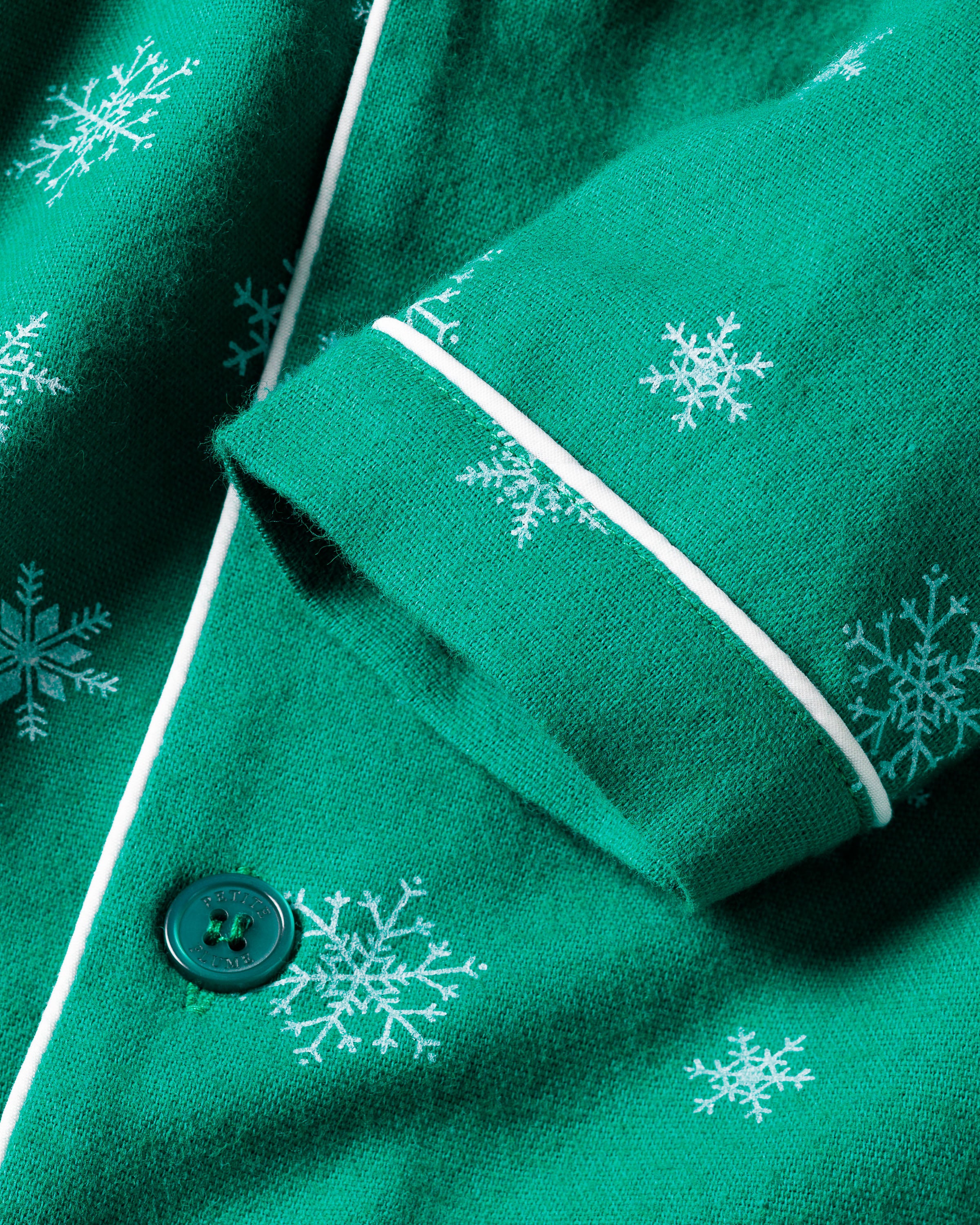 Close-up of the Kids Flannel Pajama Set in Emerald Wonderland by Petite Plume, showing green fabric with white snowflake patterns. The design includes white piping and a visible green button, ideal for moisture-wicking twill projects.