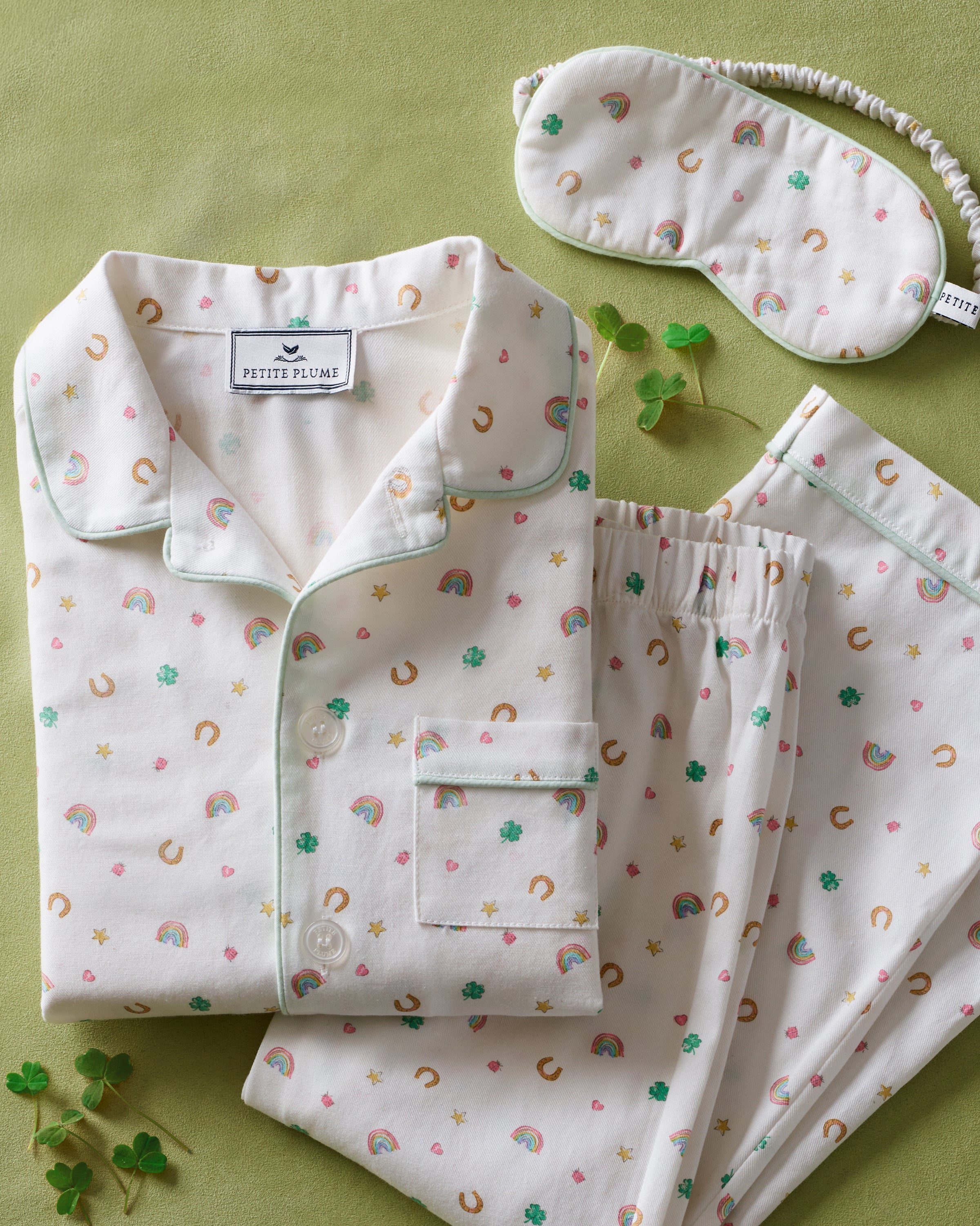 A young girl with long brown hair wears the Kid's Twill Sleep Mask in Good Luck Charms and a matching pajama top featuring rainbows, horseshoes, and clovers, standing against a neutral background.