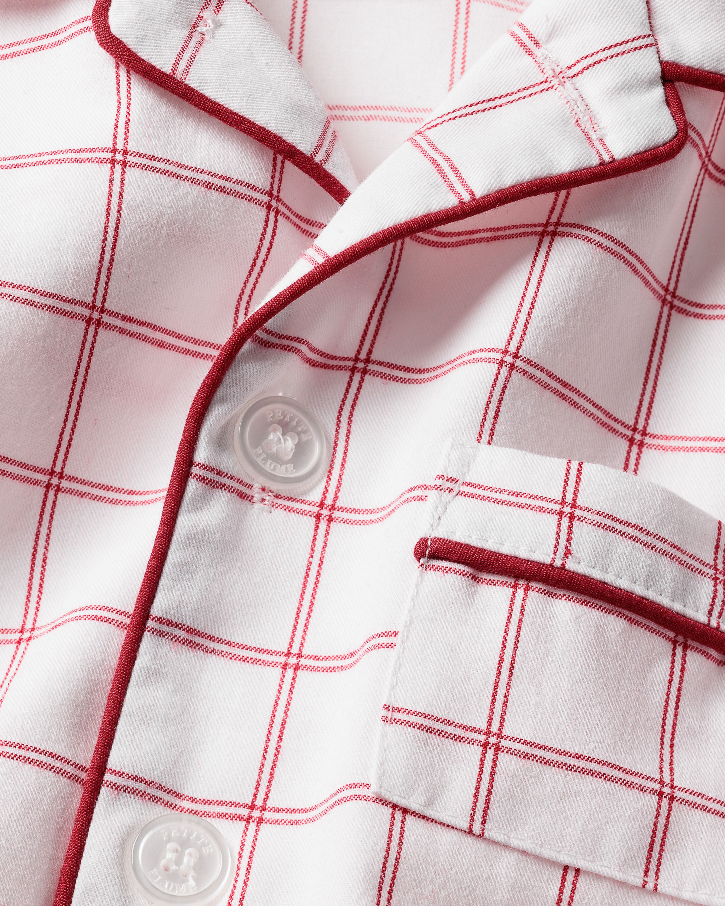 Close-up of the Kids Twill Pajama Set in Garnet Tattersall by Petite Plume, showcasing a white fabric with garnet-red checks and red-trimmed collar and pocket. It features two visible white buttons, highlighting its texture and stitching in chemical-free flame-retardant material.