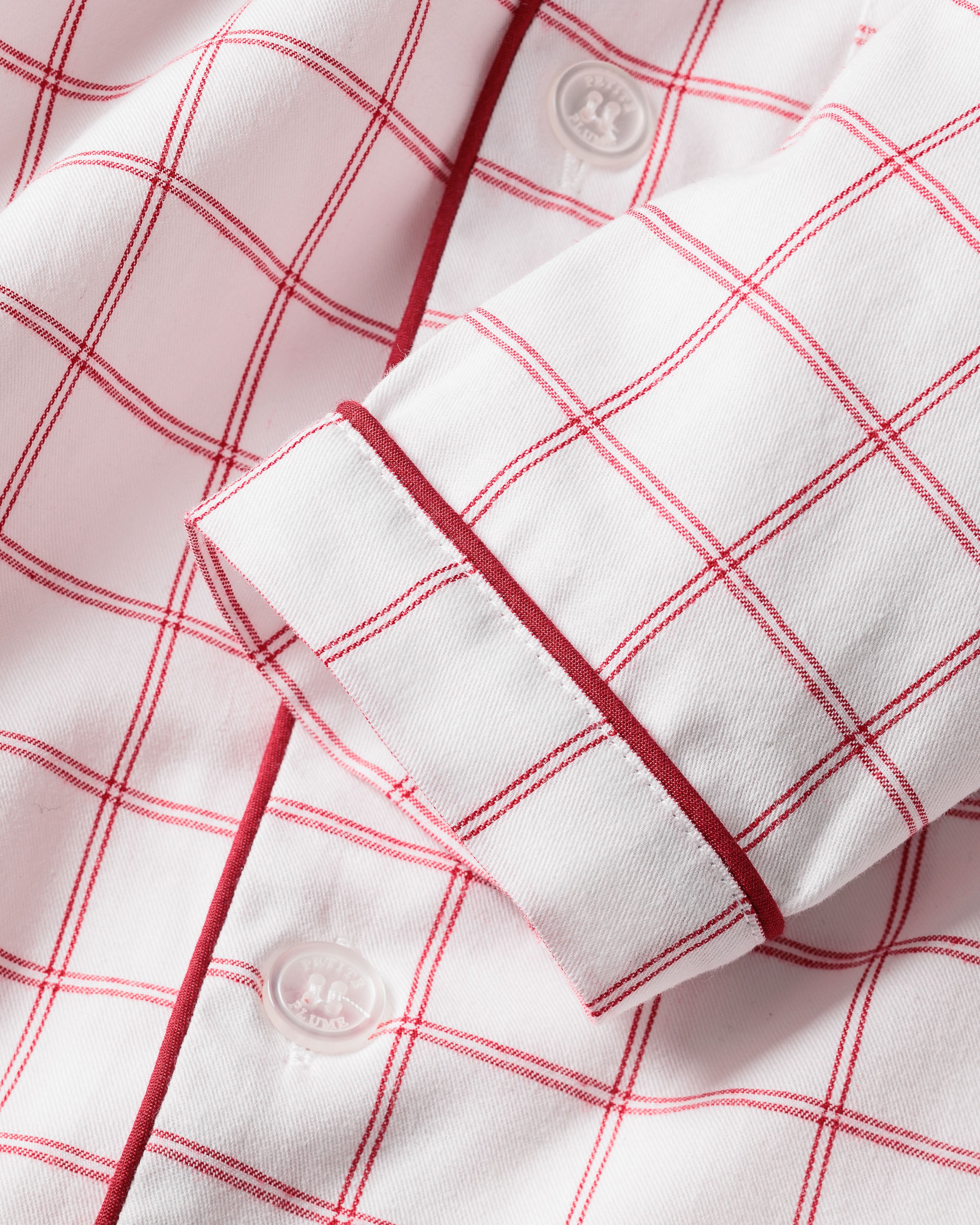 Close-up of the Kids Twill Pajama Set in Garnet Tattersall by Petite Plume, featuring a garnet-red grid pattern on white fabric. The design includes a buttoned cuff with a finely stitched red border and matching white buttons for a neat appearance.