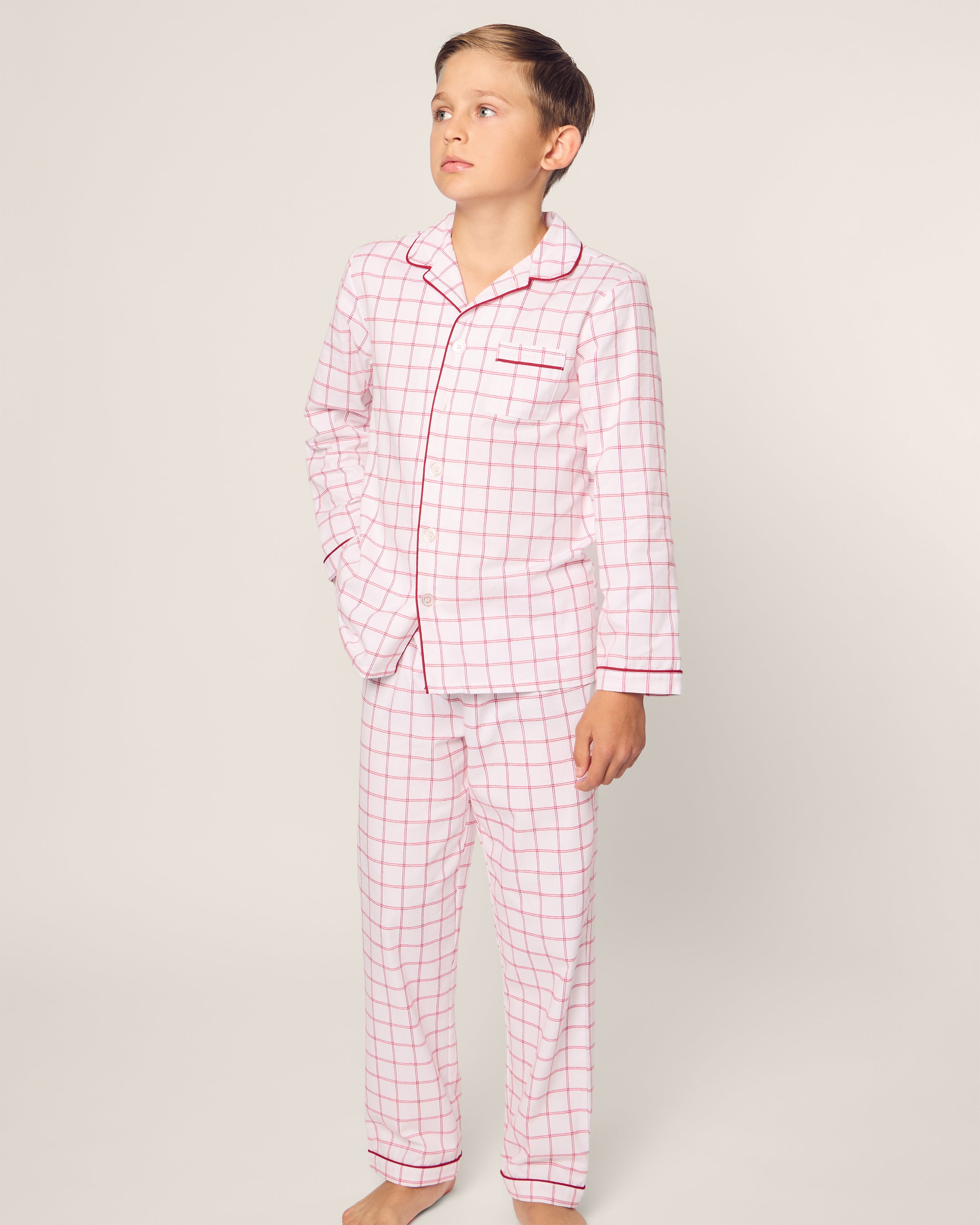 A young child models the Kids Twill Pajama Set in Garnet Tattersall by Petite Plume. These white pajamas feature a garnet-red pattern, red piping, pocket, collar, and are made from moisture-wicking twill for comfort. The child stands barefoot against a neutral backdrop.