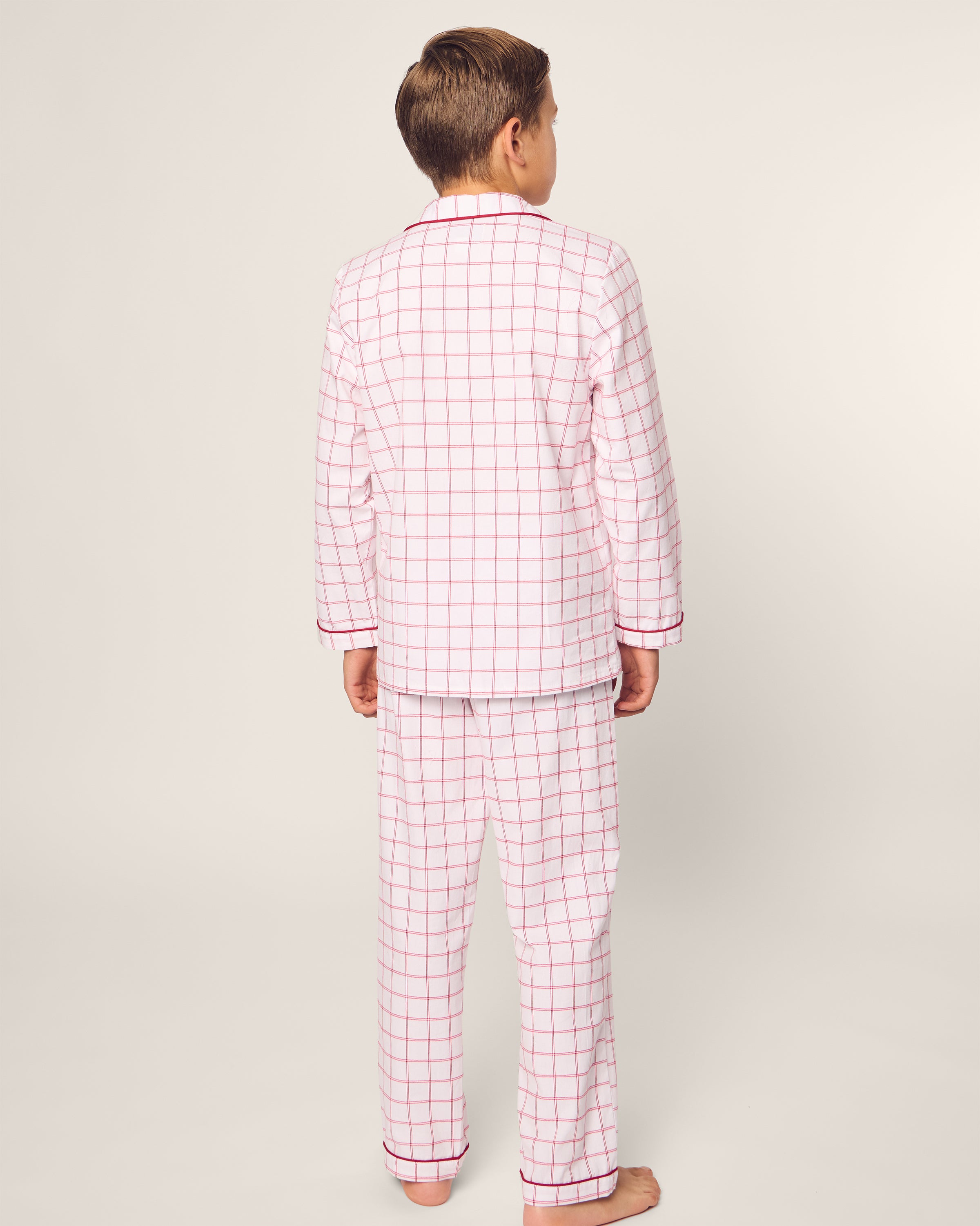 A child faces away from the camera in Petite Plumes Kids Twill Pajama Set in Garnet Tattersall, showcasing a white and pink grid pattern. The moisture-wicking twill weave includes a collared, long-sleeve top and loose-fitting pants against a plain, light-colored background.