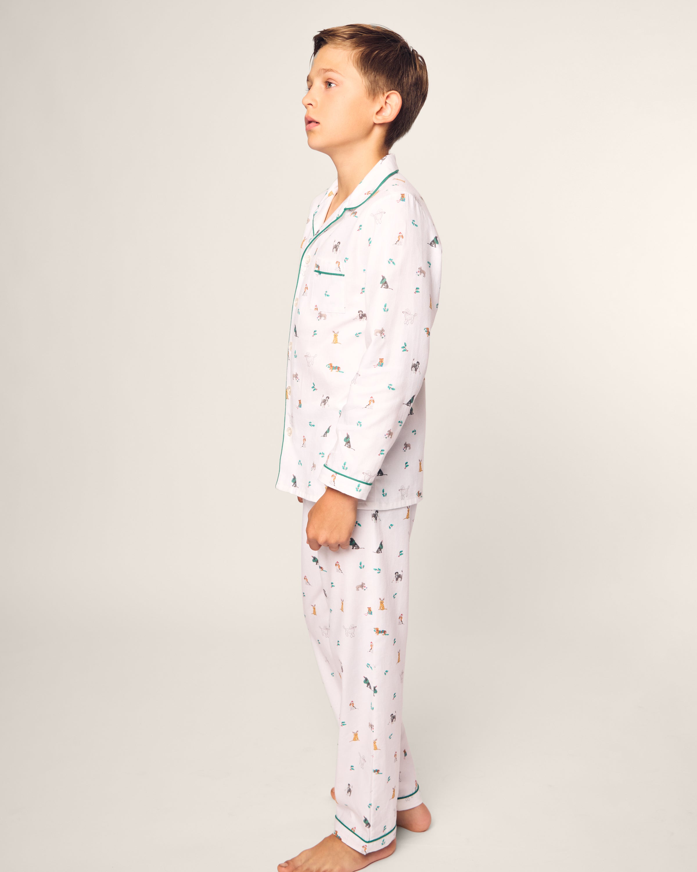 A boy stands sideways in Petite Plumes Kids Twill Pajama Set in Jingle Paws, crafted from chemical-free flame-retardant fabric, featuring colorful animal prints against a plain background.