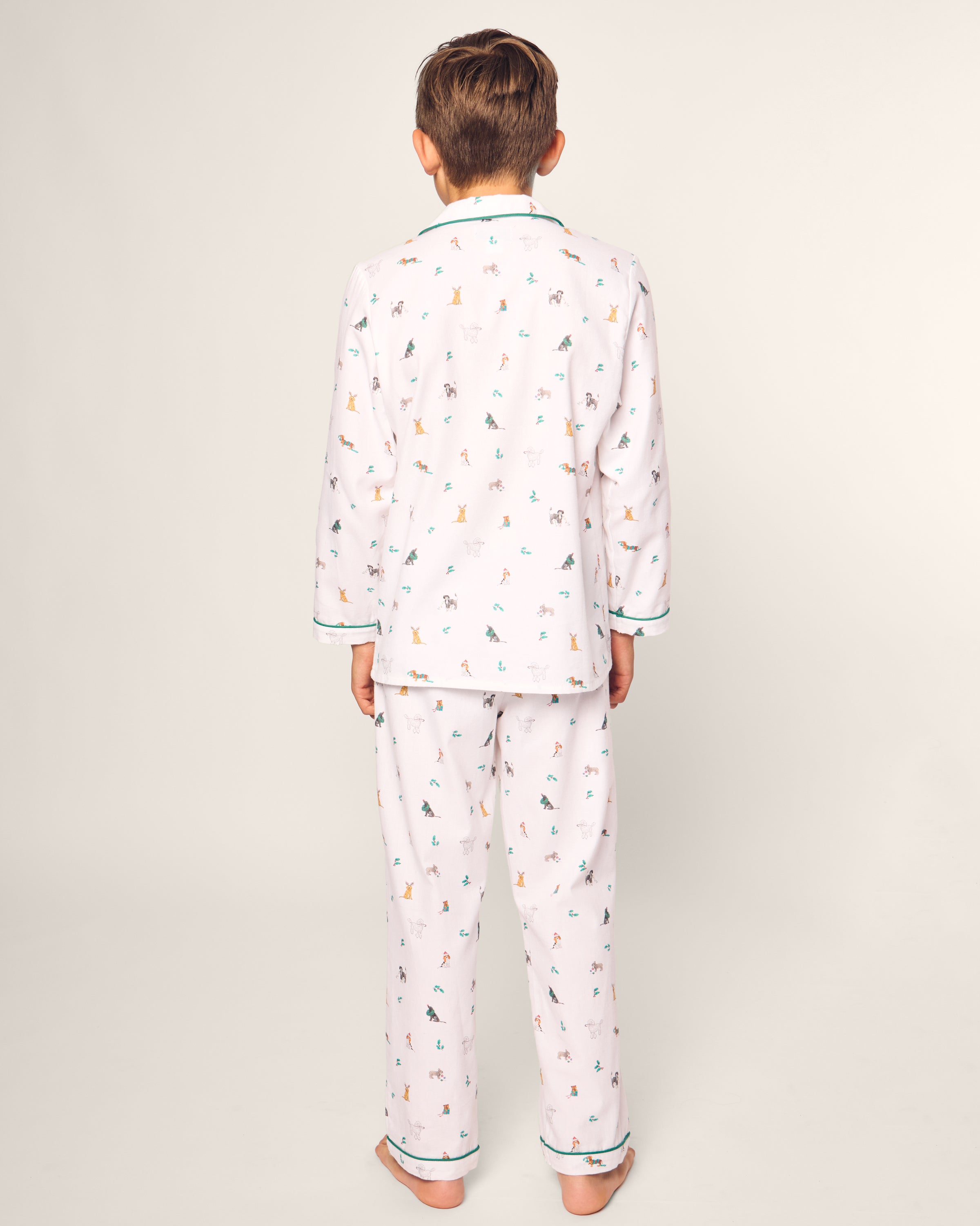 A child stands barefoot facing away, dressed in Petite Plumes Kids Twill Pajama Set in Jingle Paws. The luxury sleepwear features a playful animal print on chemical-free flame-retardant fabric. The button-down top and long pants have contrasting trim, set against a plain background for emphasis.