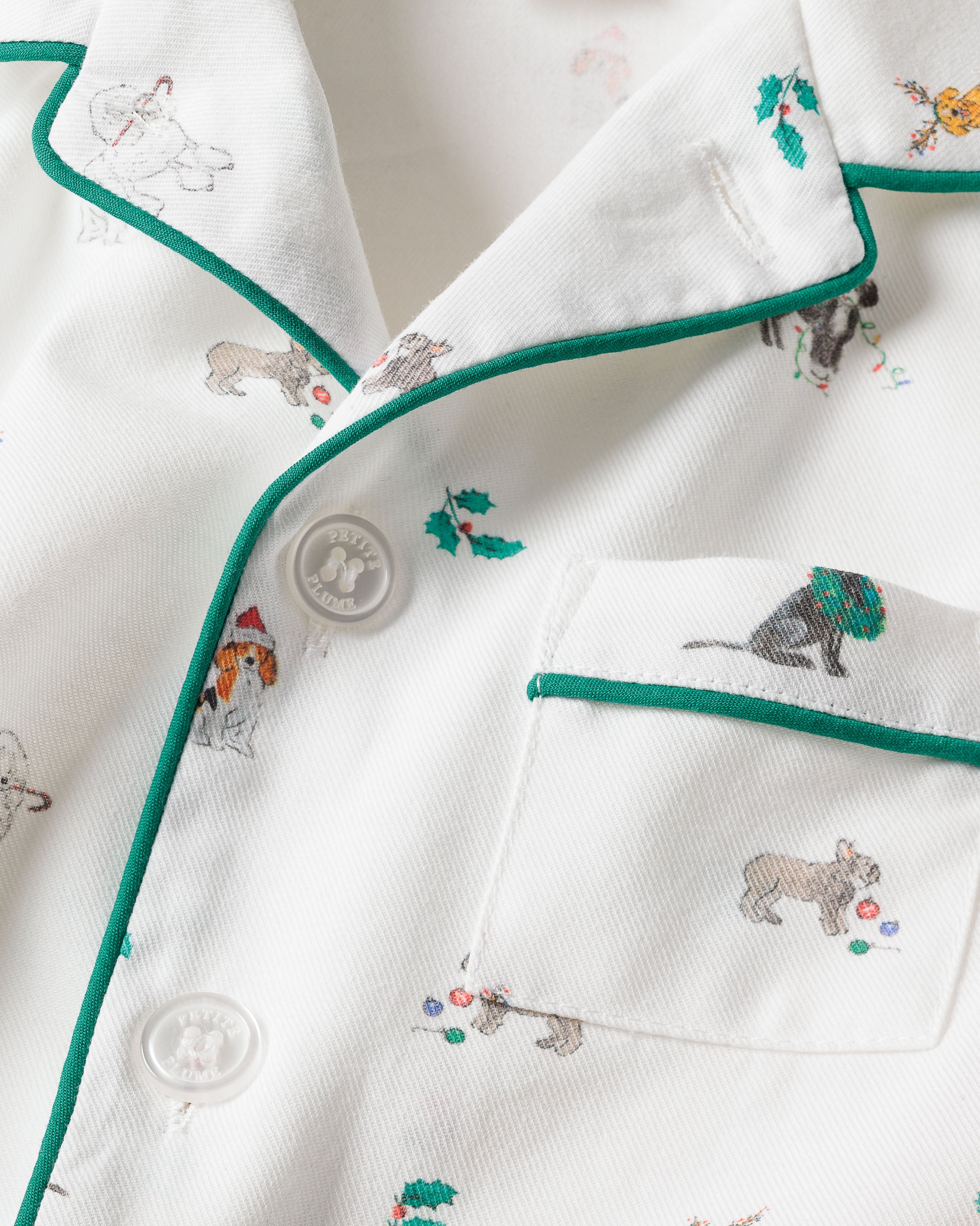 The Kids Twill Pajama Set in Jingle Paws by Petite Plume features a white top with green piping, button-up front, and pocket. Adorned with playful dog illustrations in festive attire and tiny holly patterns, its made from chemical-free flame-retardant fabric for peace of mind.