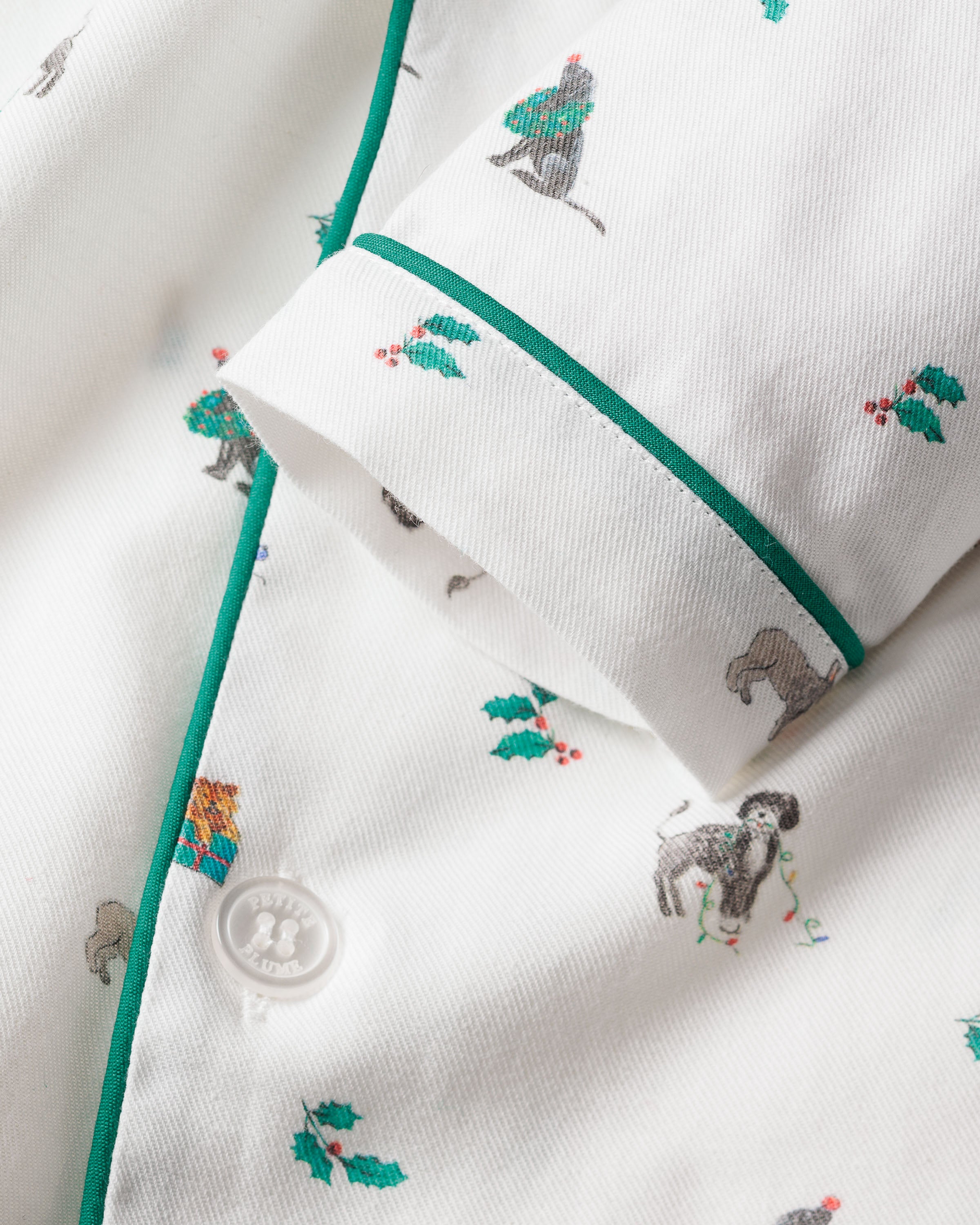 The Kids Twill Pajama Set in Jingle Paws by Petite Plume features a white fabric adorned with festive animals, holly leaves, and bright green piping. A charming white button adds a touch of luxury, making it perfect for childrens pajamas.