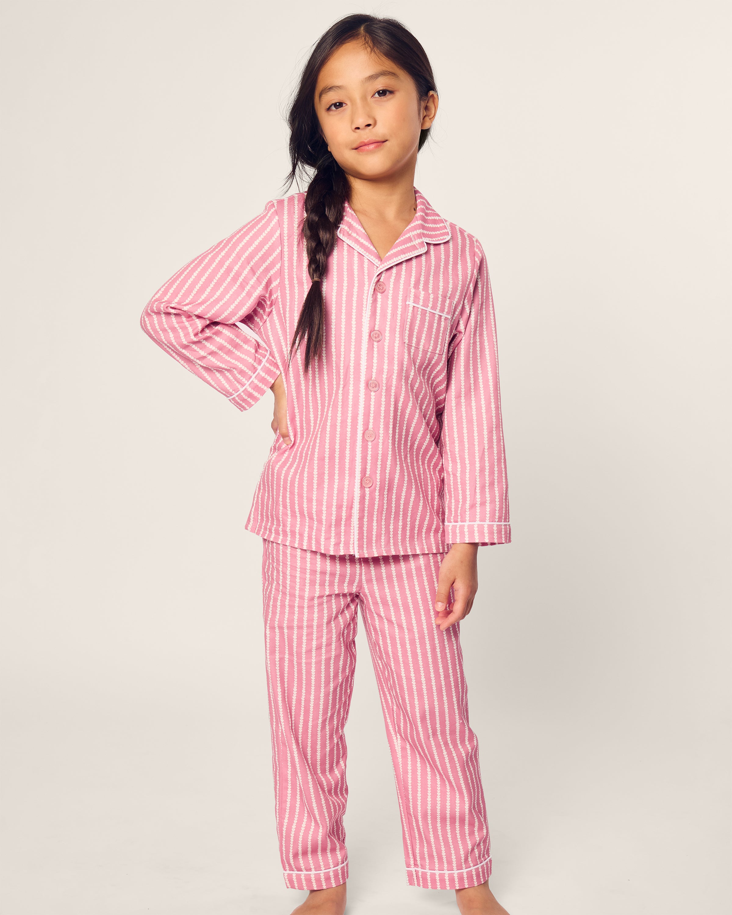 A young girl stands confidently in Petite Plumes Kids Twill Pajama in Love Lines, featuring flame retardant fiber. Shes wearing pink and white striped pajamas with a collar and long sleeves. Her long dark hair is styled in a side braid as she poses with one hand on her hip against a plain background.