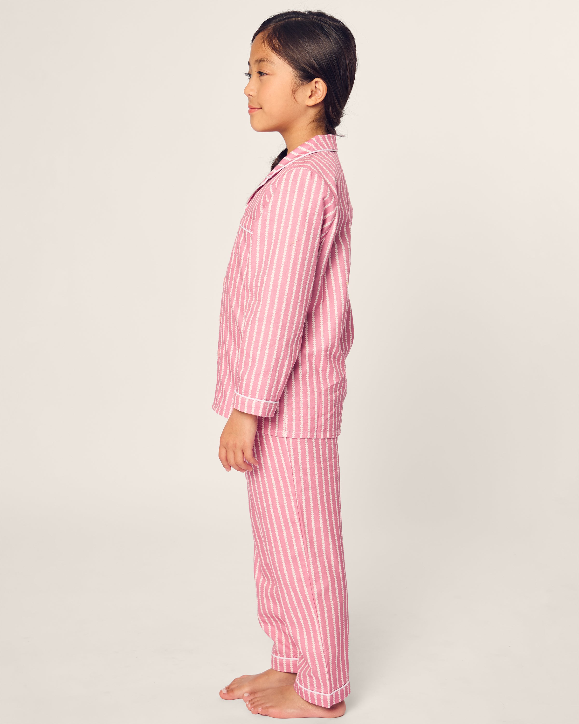 Side profile of a child standing barefoot on a plain, light background, wearing Petite Plumes Kids Twill Pajama in Love Lines with pink and white stripes.