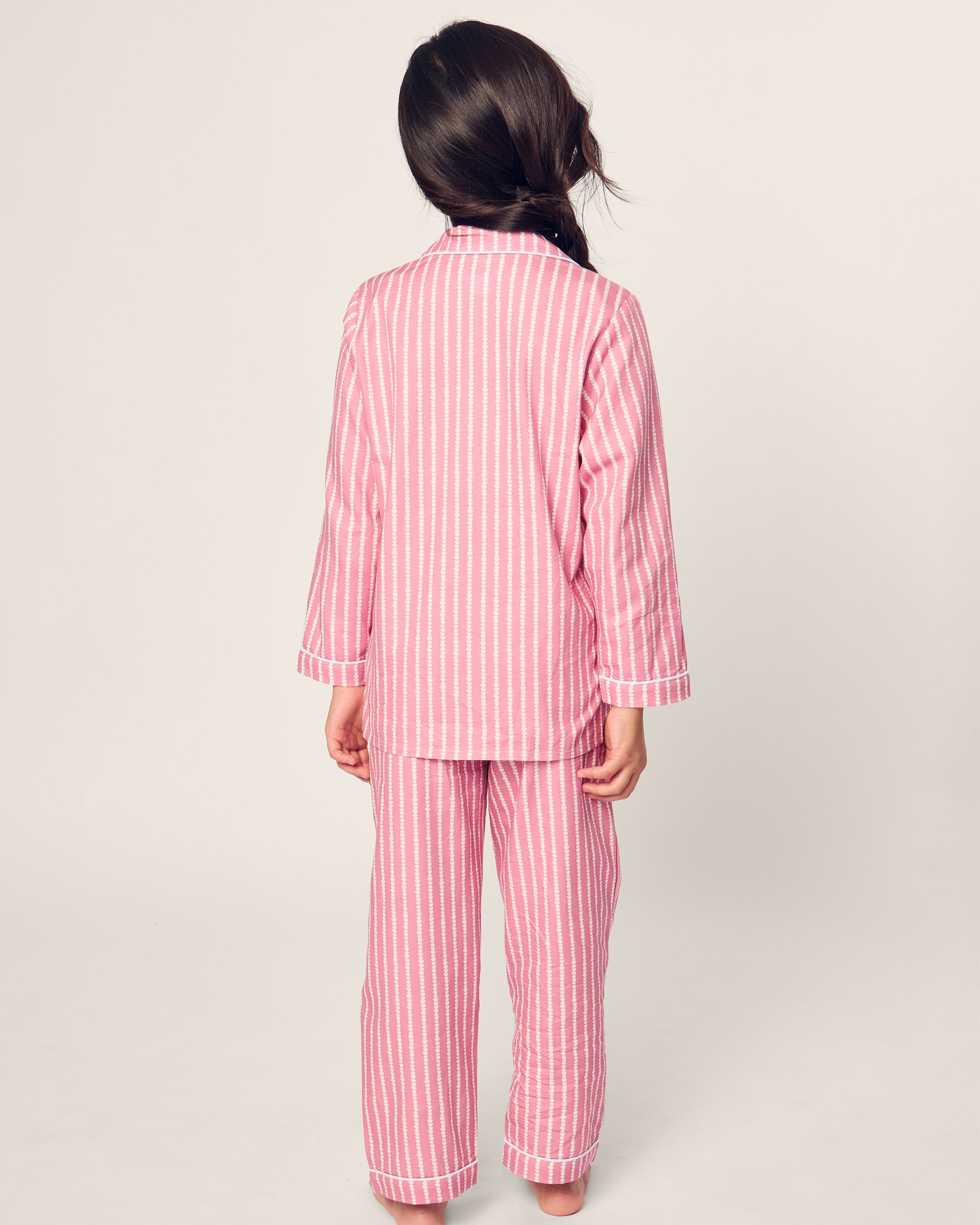 A child in Petite Plumes Kids Twill Pajama in Love Lines, made of cozy yarn-dyed cotton, stands with their back to the camera. The comfy long sleeves and pants are shown as a neat braid cascades over the left shoulder against an off-white backdrop.