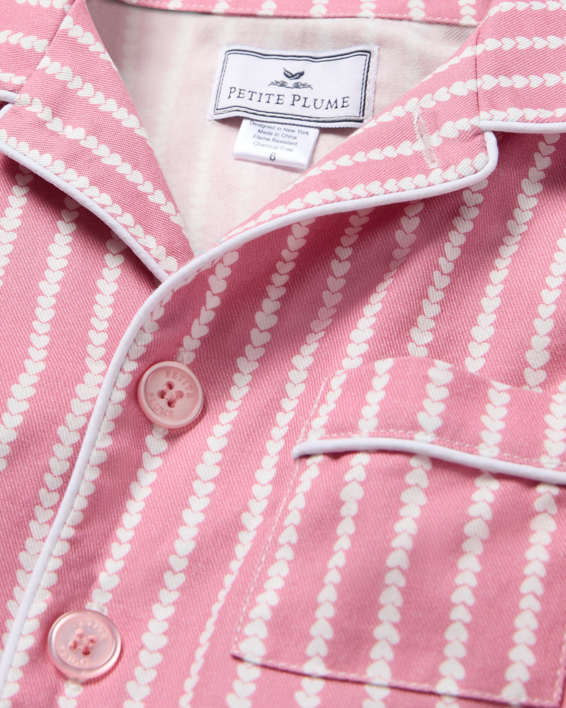 Close-up of Petite Plumes Kids Twill Pajama Top in Love Lines, featuring pink fabric with white heart-striped patterns, white piping, two buttons, and a chest pocket. Crafted from yarn-dyed cotton as part of a matching set.