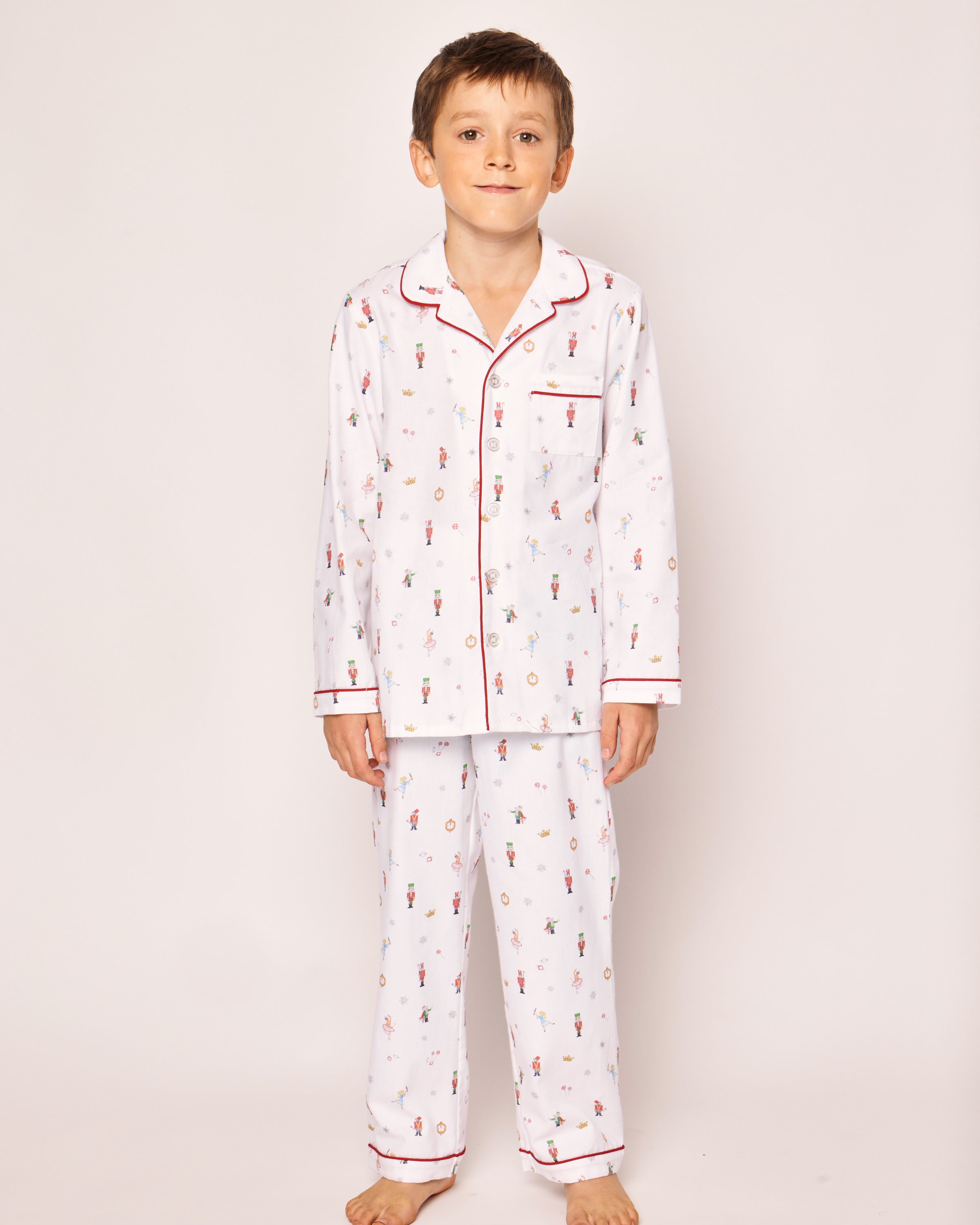 A young boy stands barefoot in Petite Plumes Kids Twill Pajama Set, A Night at the Nutcracker, featuring a festive pattern and red piping. The chemical-free sleepwear includes a long-sleeve button-up shirt and matching pants. He is against a plain, light-colored background.