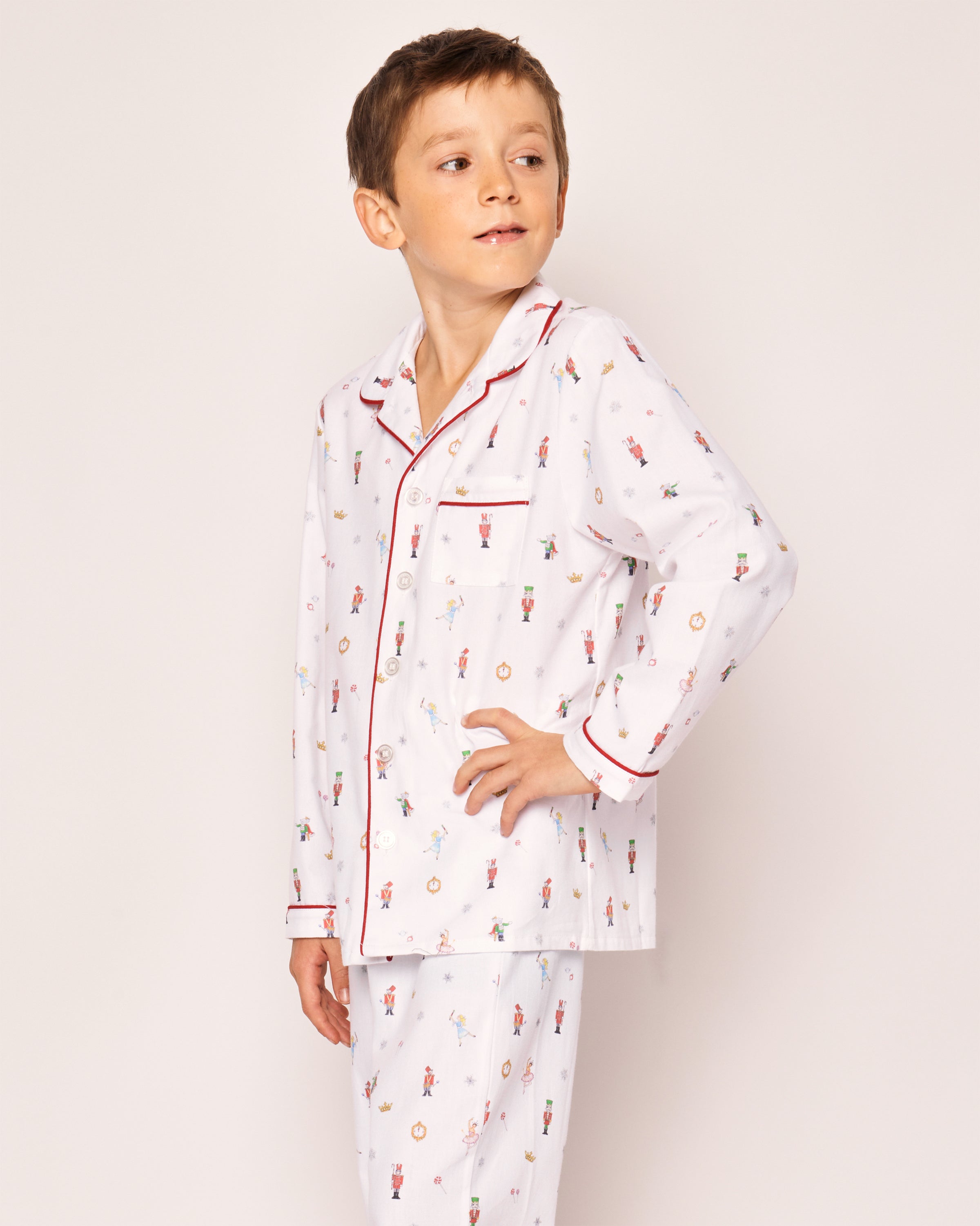 A young boy wears Petite Plumes Kids Twill Pajama Set in A Night at the Nutcracker, showcasing colorful prints with red piping on the collar and pocket. These chemical-free pajamas offer safety and comfort as he looks to his right against a plain background.
