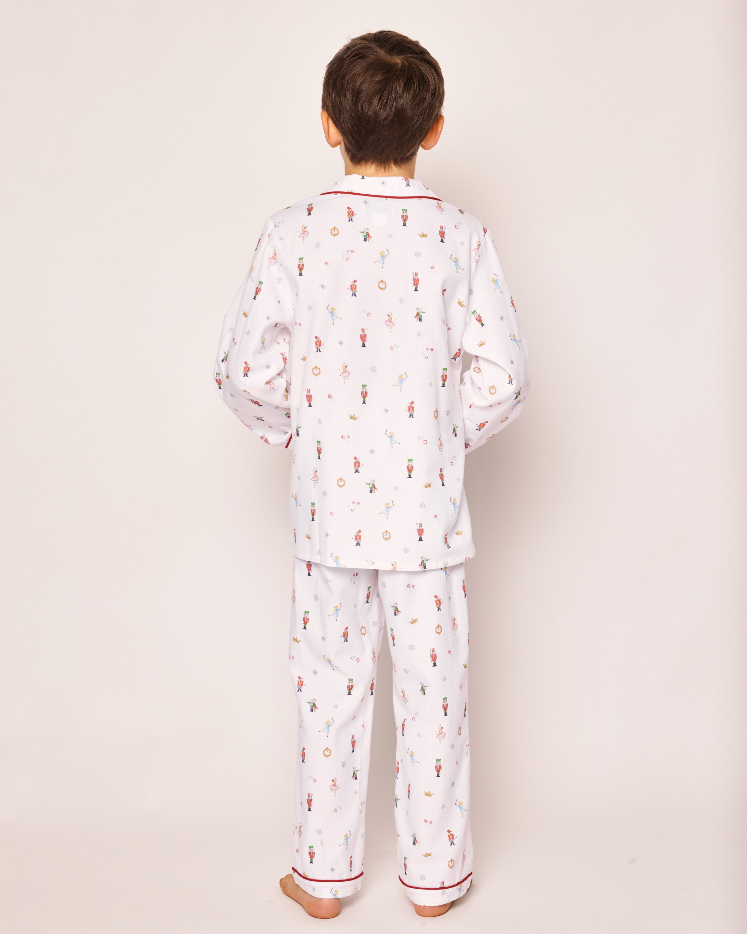 A child stands barefoot against a plain background wearing Petite Plumes Kids Twill Pajama Set in A Night at the Nutcracker, featuring relaxed-fit pajamas with colorful prints on chemical-free material for an enchanting nights rest.
