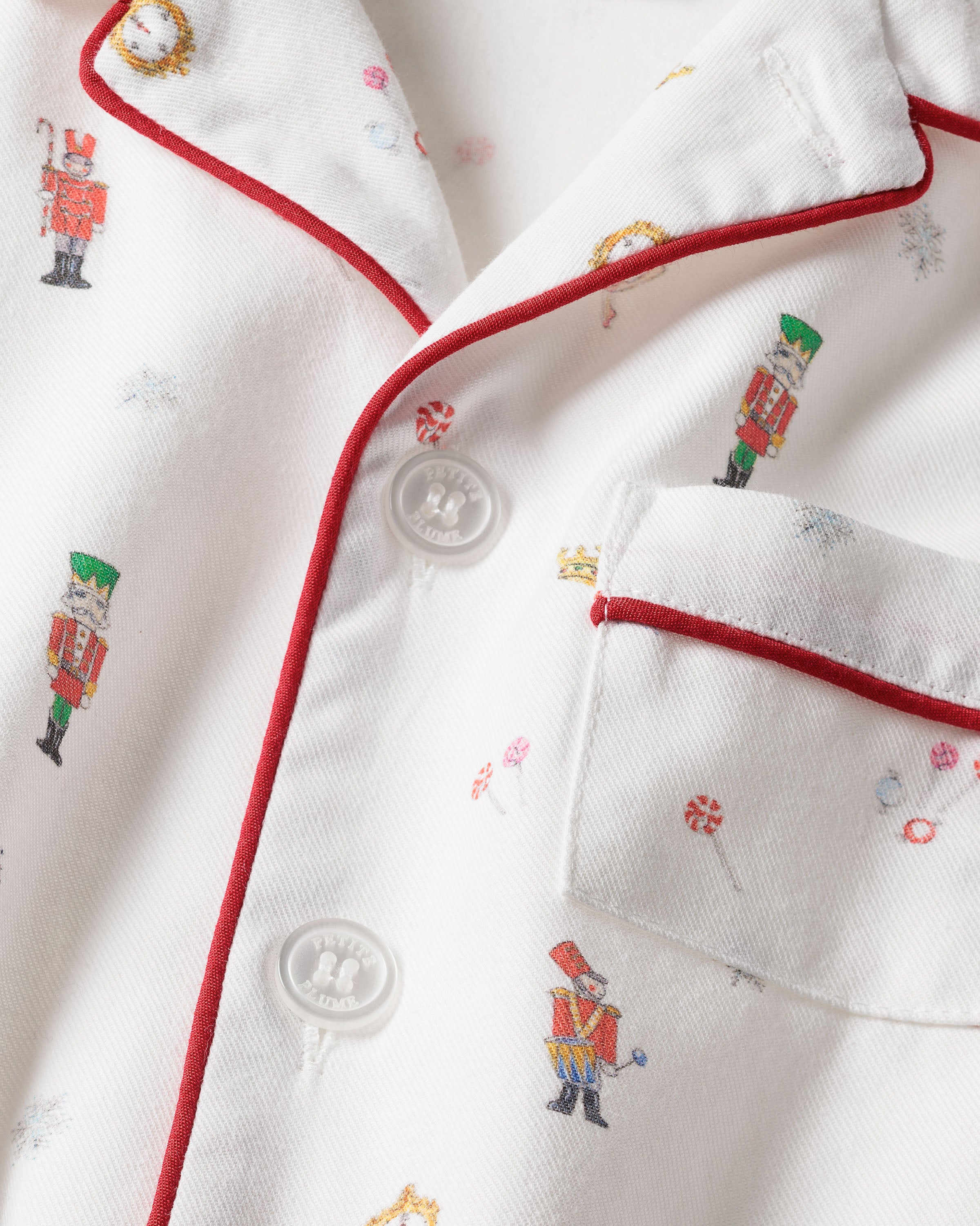 A close-up of the fabric reveals small illustrations of toy soldiers, candy, and drums on a white background. Petite Plumes Kids Twill Pajama Set in A Night at the Nutcracker has red piping along the collar and pocket, white buttons, and is made with childrens flame-retardant fabric.