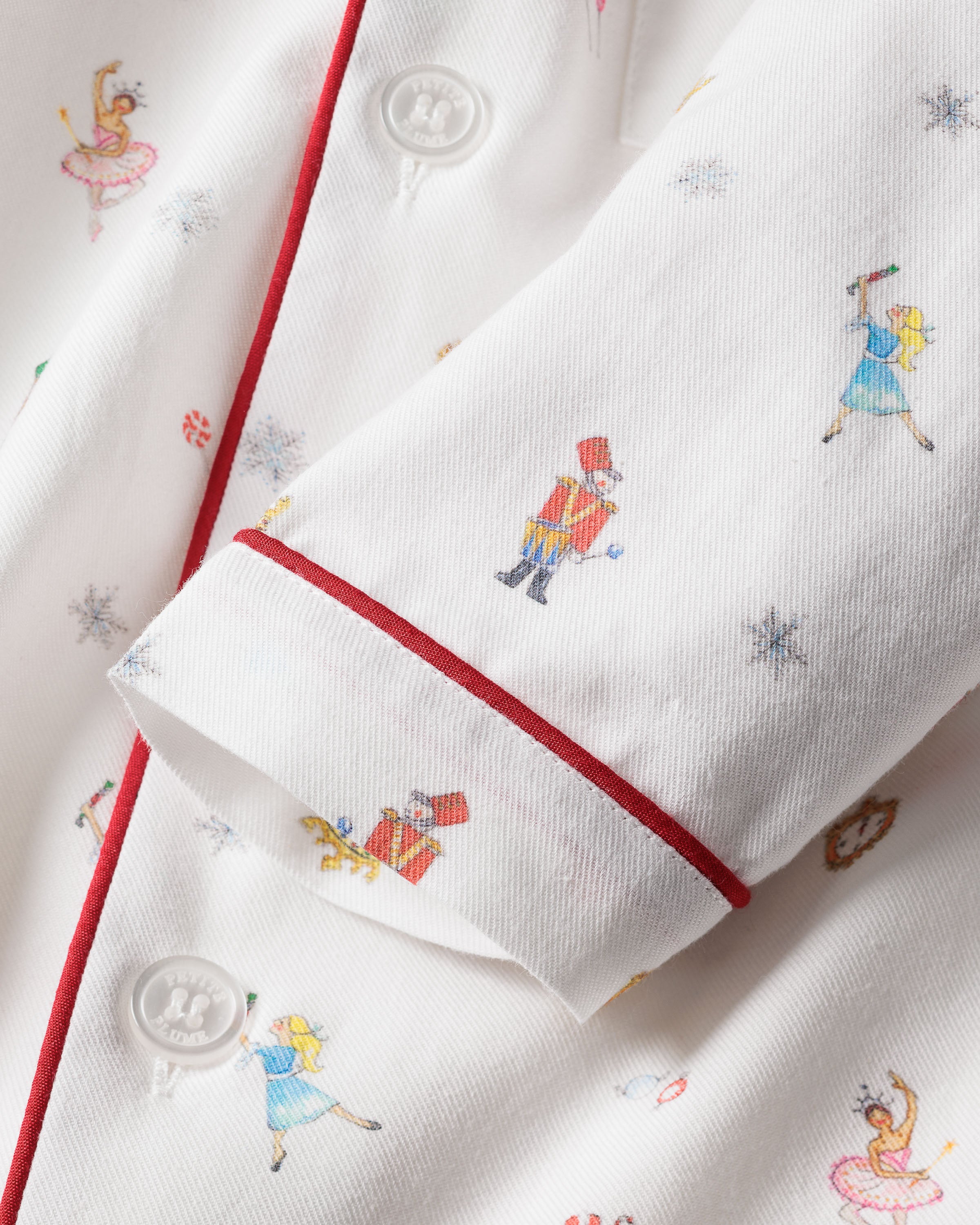 Close-up of Petite Plumes Kids Twill Pajama Set in A Night at the Nutcracker shows white fabric with red piping, printed figures like soldiers and dancers, hinting at Nutcracker-inspired pajamas for children. Perfect chemical-free sleepwear featuring sleeve and button details for little dreamers.