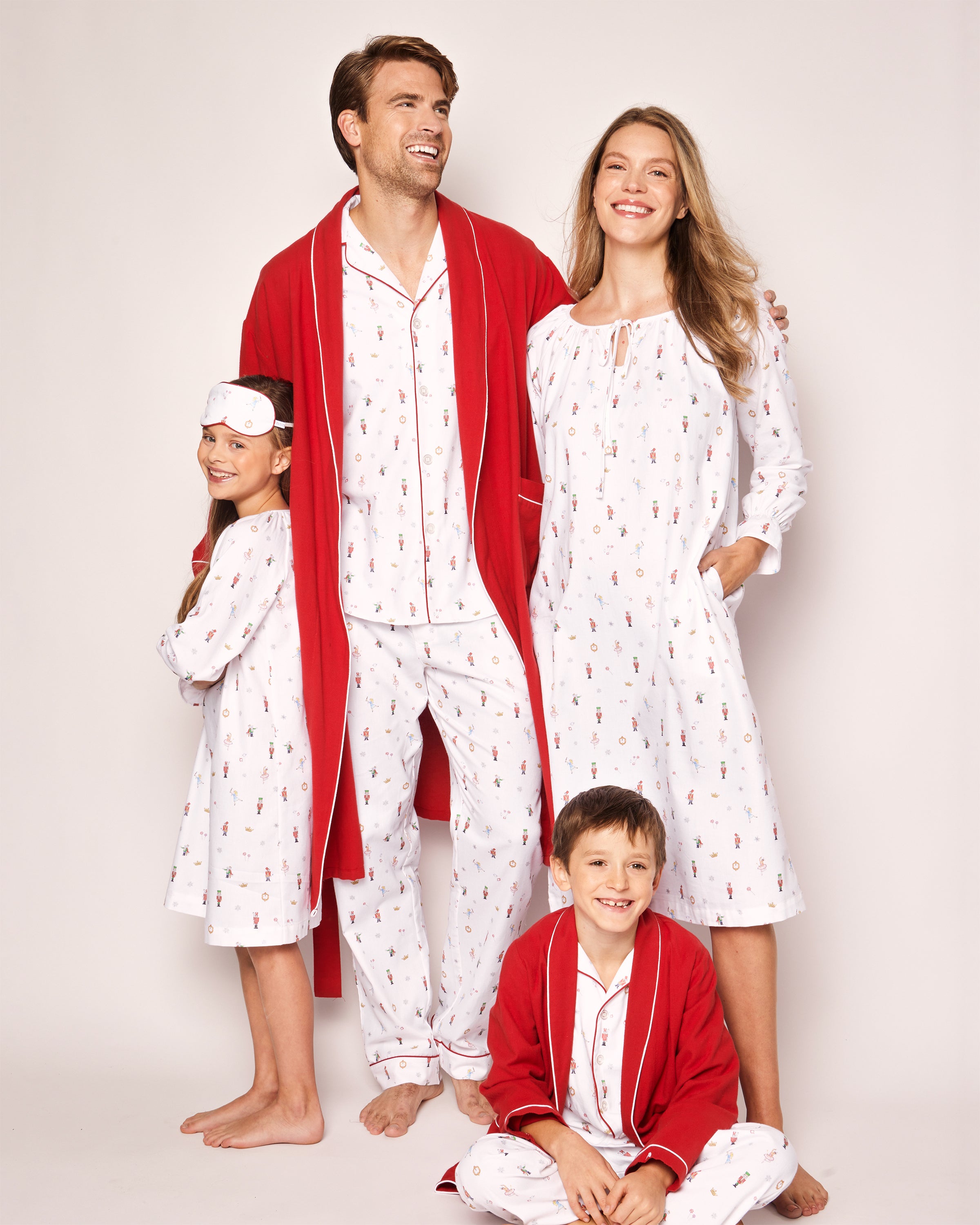The family smiles warmly in their Petite Plume sleepwear: the son wears a Kids Twill Pajama Set in A Night at the Nutcracker, paired with his father’s red robe, while the mother and daughter sport charming nightgowns. They enjoy the comfort of chemical-free pajamas against a plain backdrop.