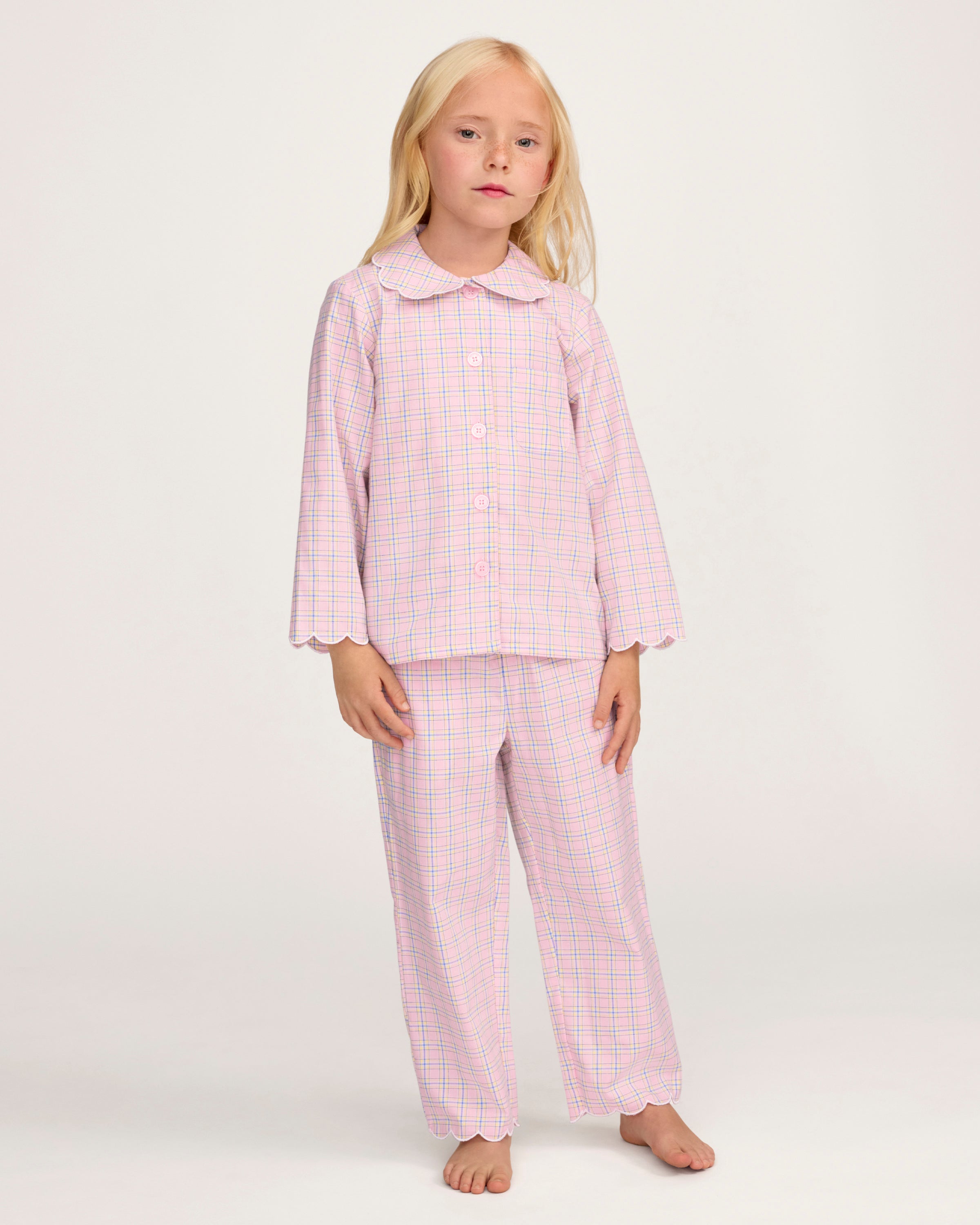 Young girl with long blonde hair wears the Kid's Twill Colette Pajama Set in Peony Plaid, featuring pink and white checks, scalloped edges, and stands barefoot against a plain light background.