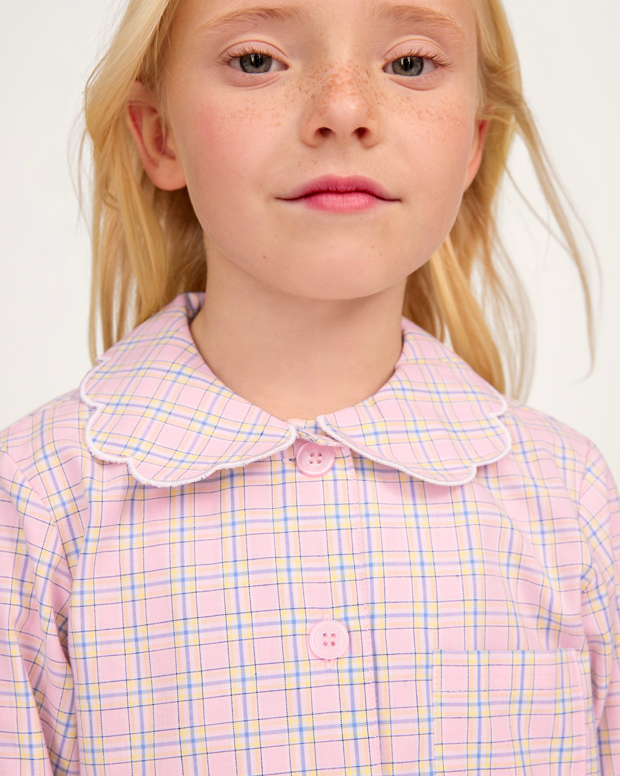 Young girl with long blonde hair wears the Kid's Twill Colette Pajama Set in Peony Plaid, featuring pink and white checks, scalloped edges, and stands barefoot against a plain light background.